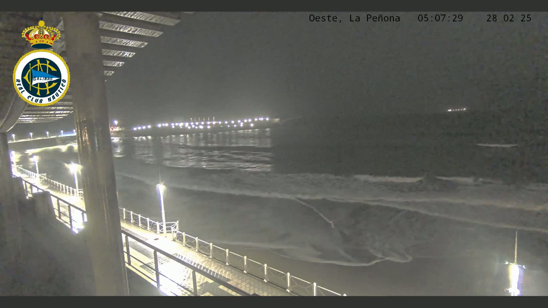 A nighttime view from a pier shows dark ocean waves lapping the shore under a dark sky, with lights illuminating the pier and a distant coastline.
