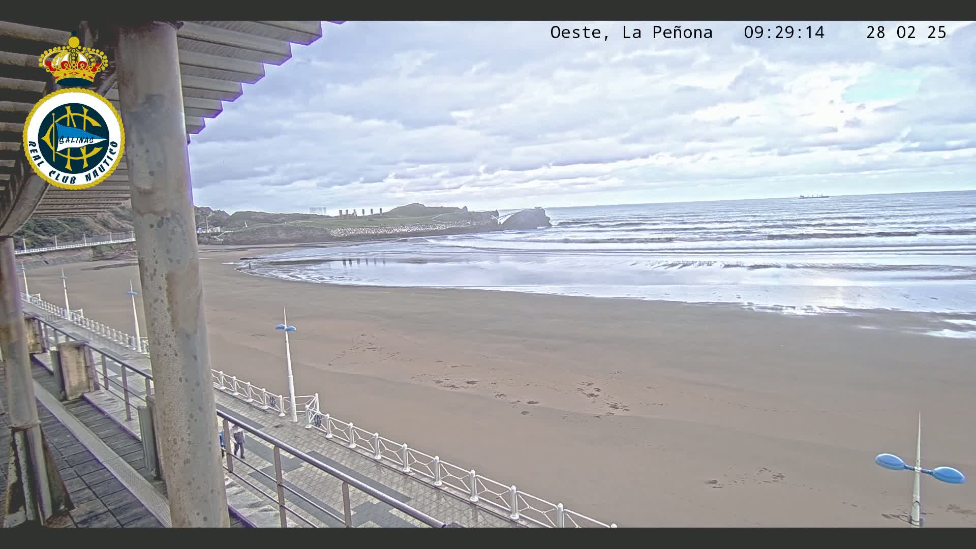 A sandy beach with moderate waves under a cloudy sky is viewed from an elevated position, showing a boardwalk and a distant rocky outcrop.