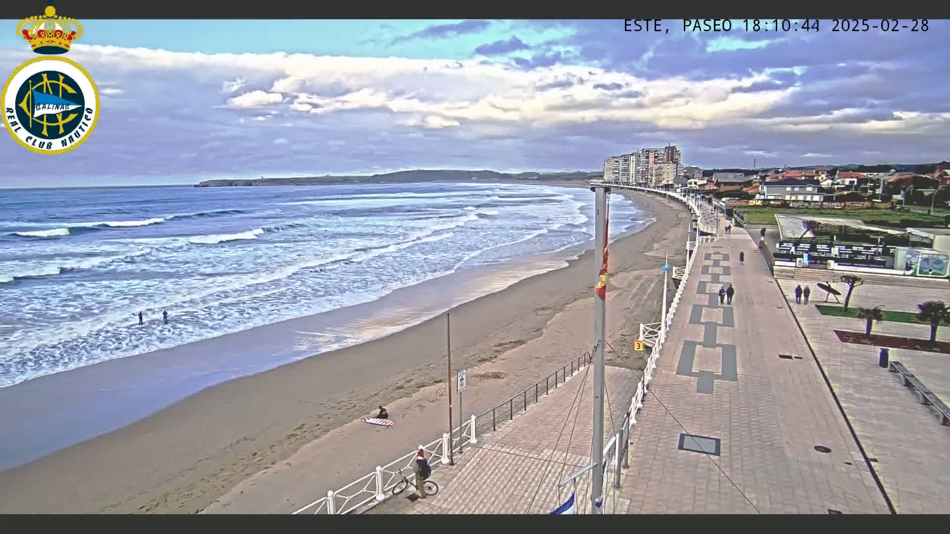 A sandy beach with moderate waves under a partly cloudy sky is next to a paved boardwalk with a few pedestrians and cyclists, bordered by buildings.