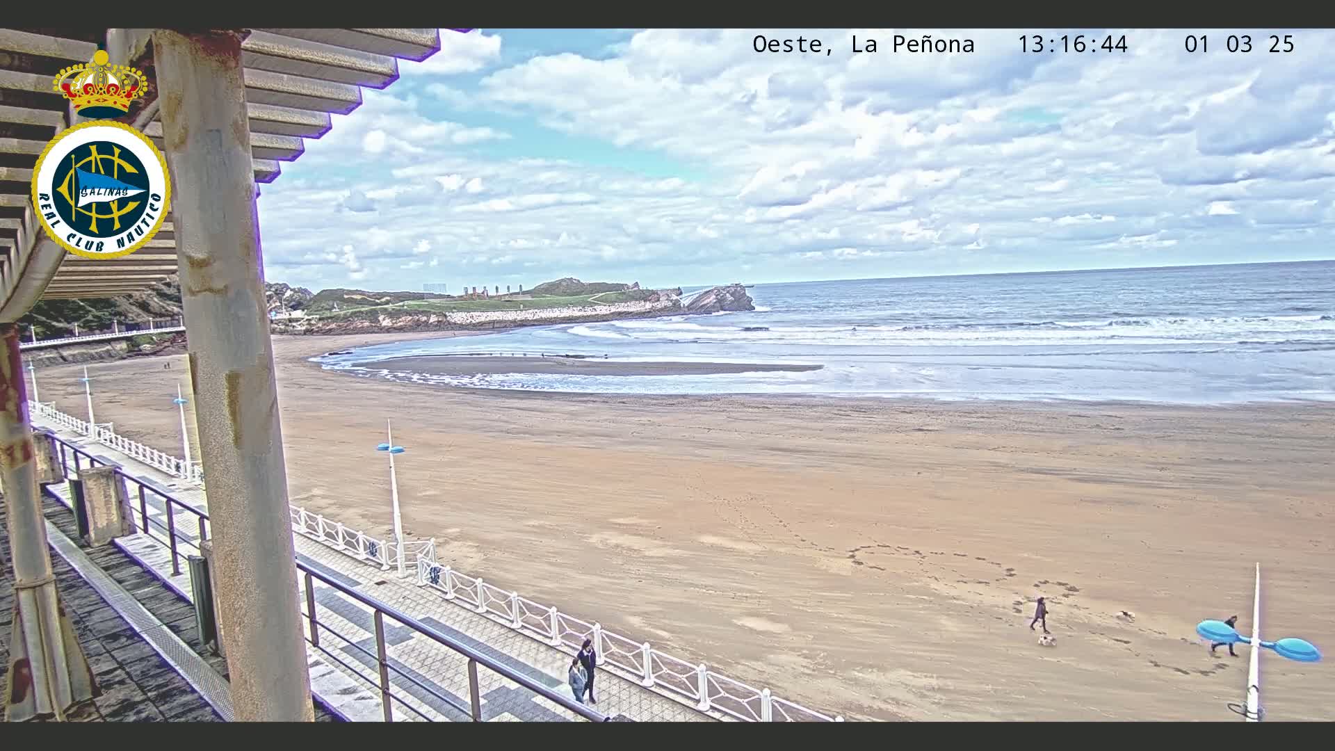 A sandy beach with a few people walking along a boardwalk, under a partly cloudy sky, and a rocky outcrop visible in the distance.