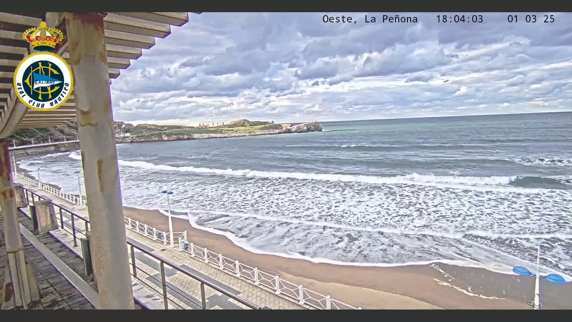 A sandy beach with moderate waves under a partly cloudy sky is viewed from a covered walkway.