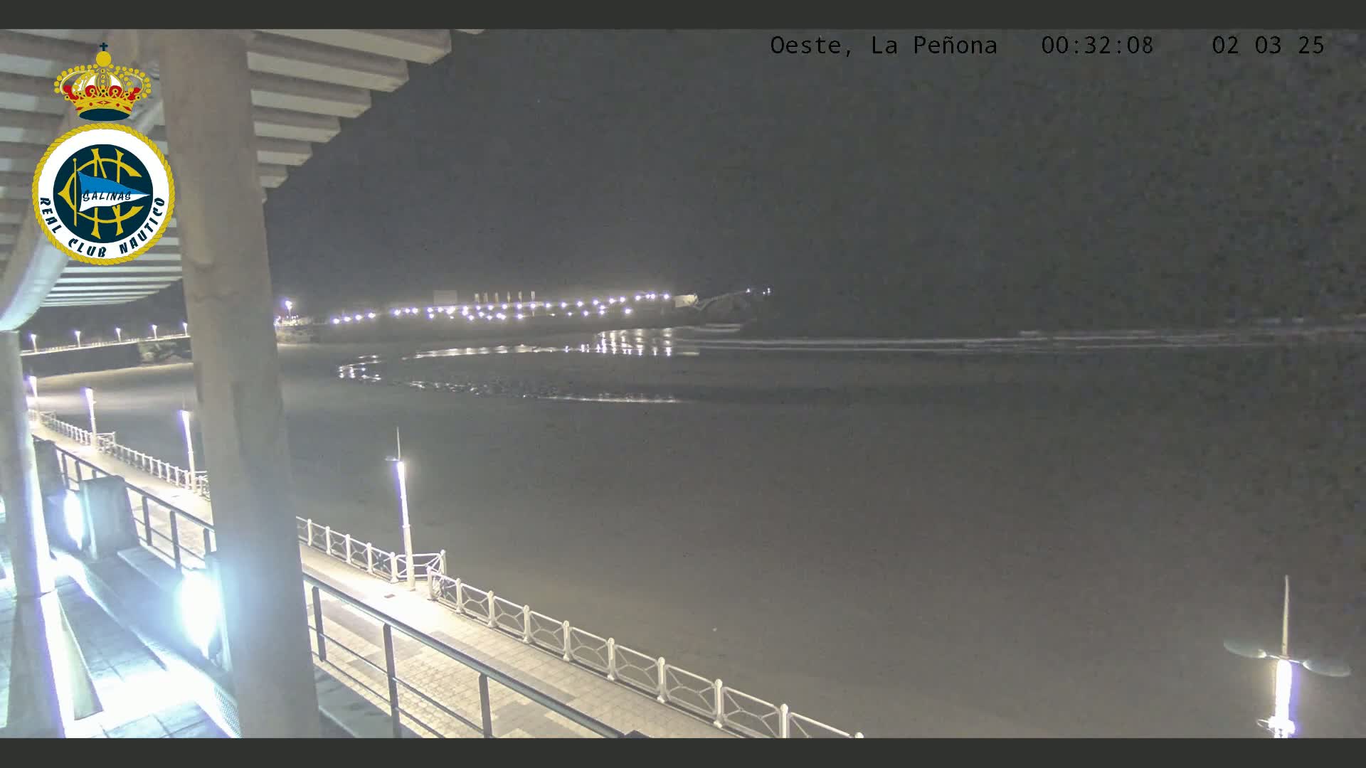 A nighttime view of a beach with calm water, a boardwalk with lights, and a building with a partially visible structure overhead.