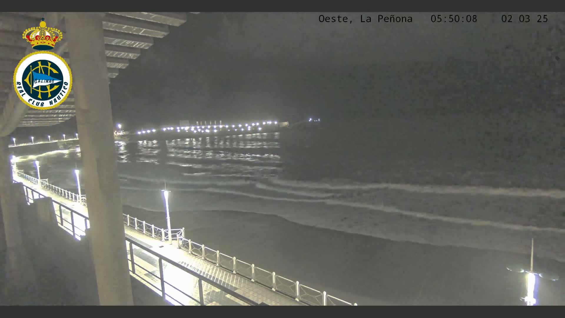 A nighttime view from a pier shows a dark ocean with small waves gently lapping the shore under a cloudy sky.
