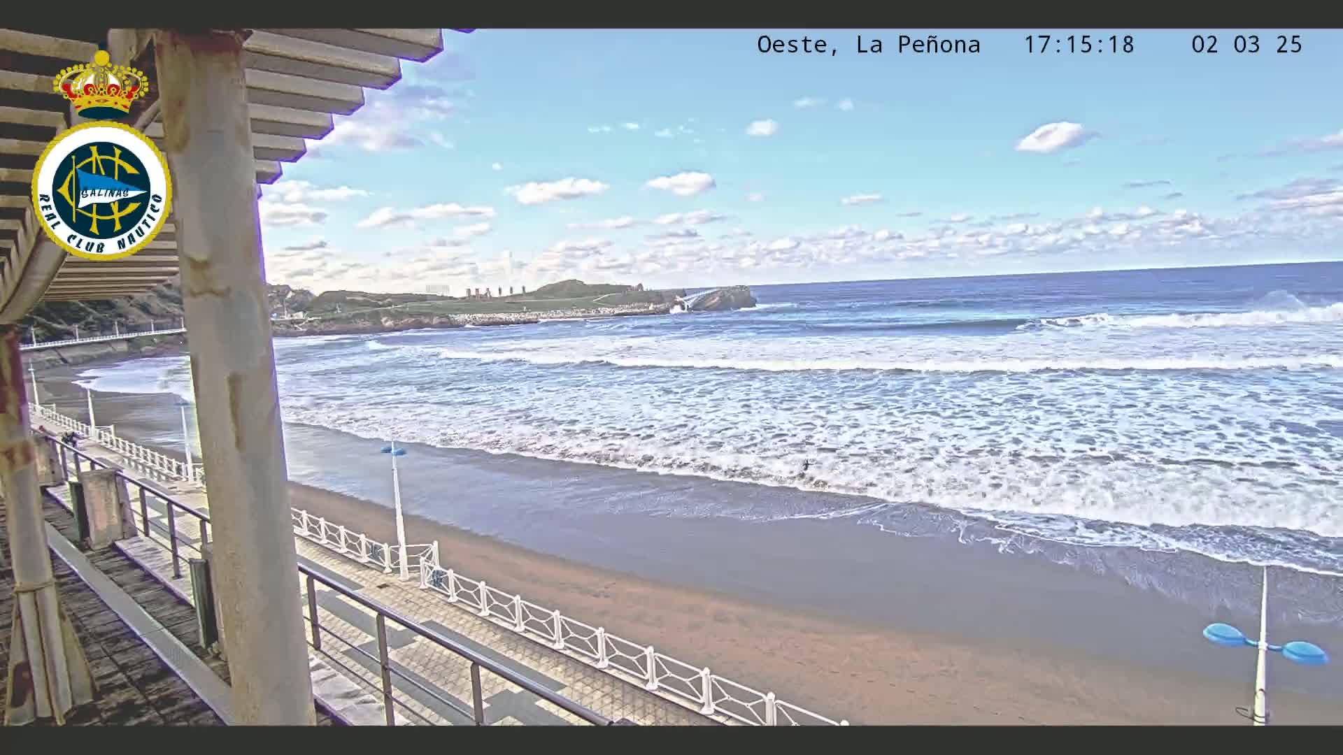 A partly cloudy day shows an ocean beach with waves rolling onto a sandy shore, viewed from a balcony with a walkway and railing.