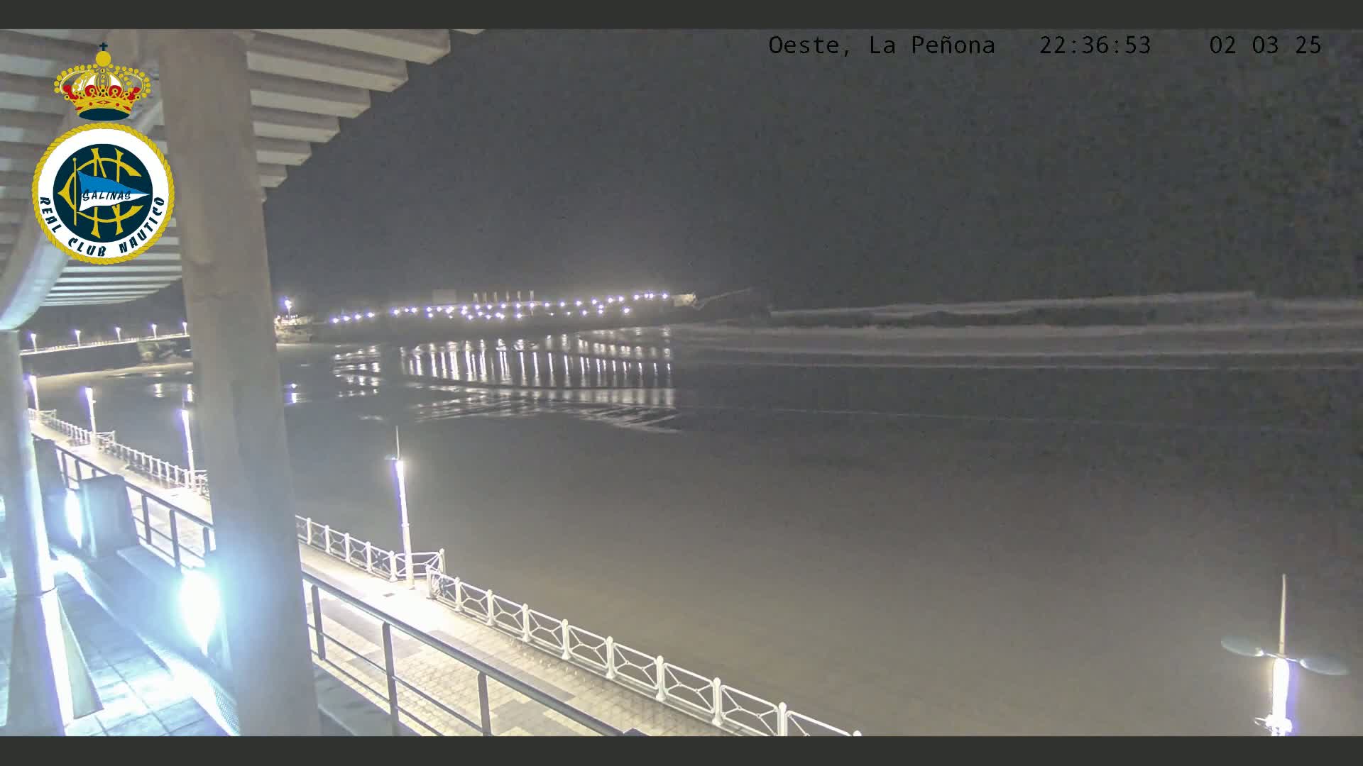 A nighttime view of a calm ocean, illuminated by lights along a pier and a shoreline.
