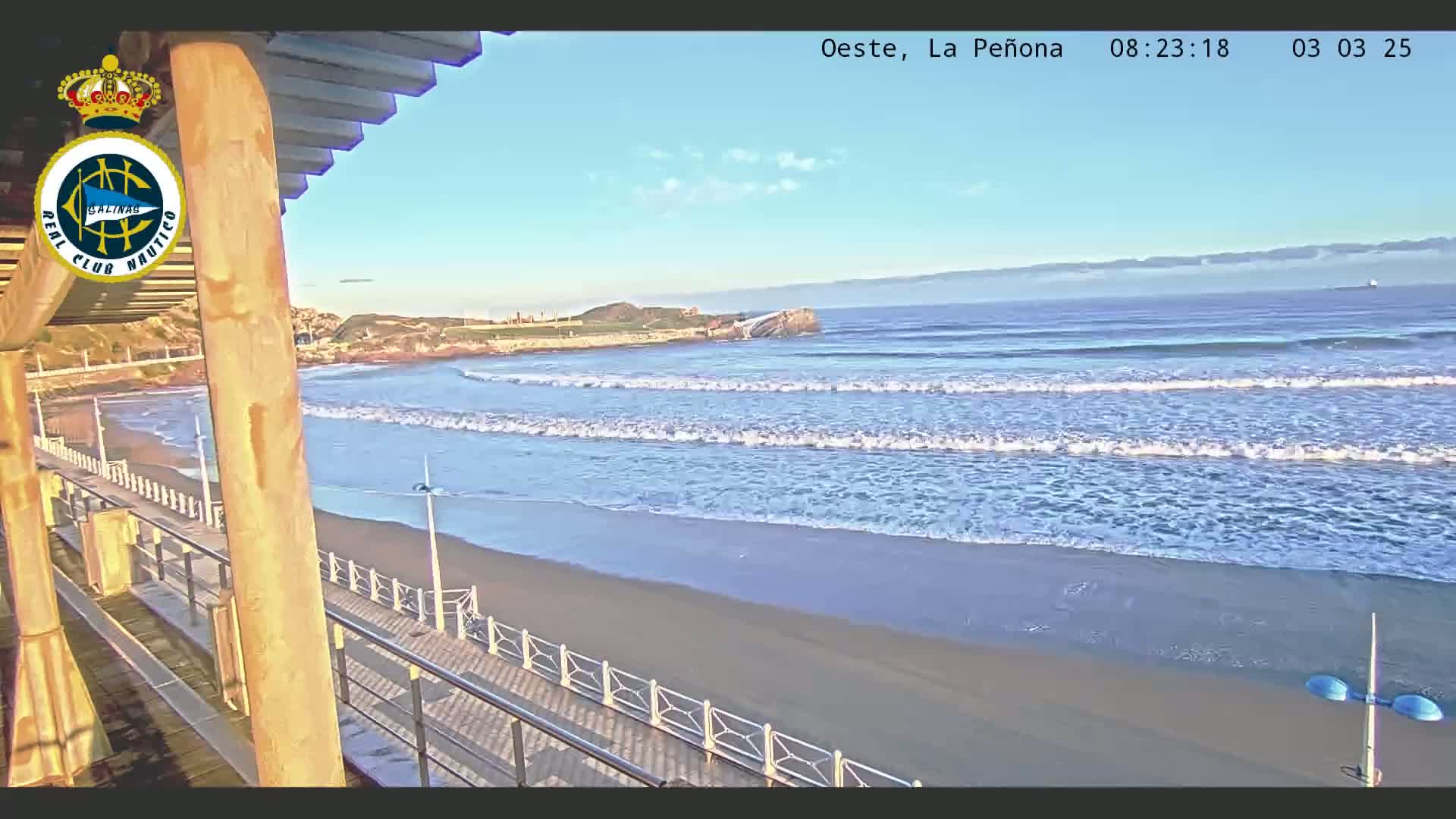 A sandy beach with gentle waves rolls onto the shore under a mostly clear sky, viewed from an elevated boardwalk.