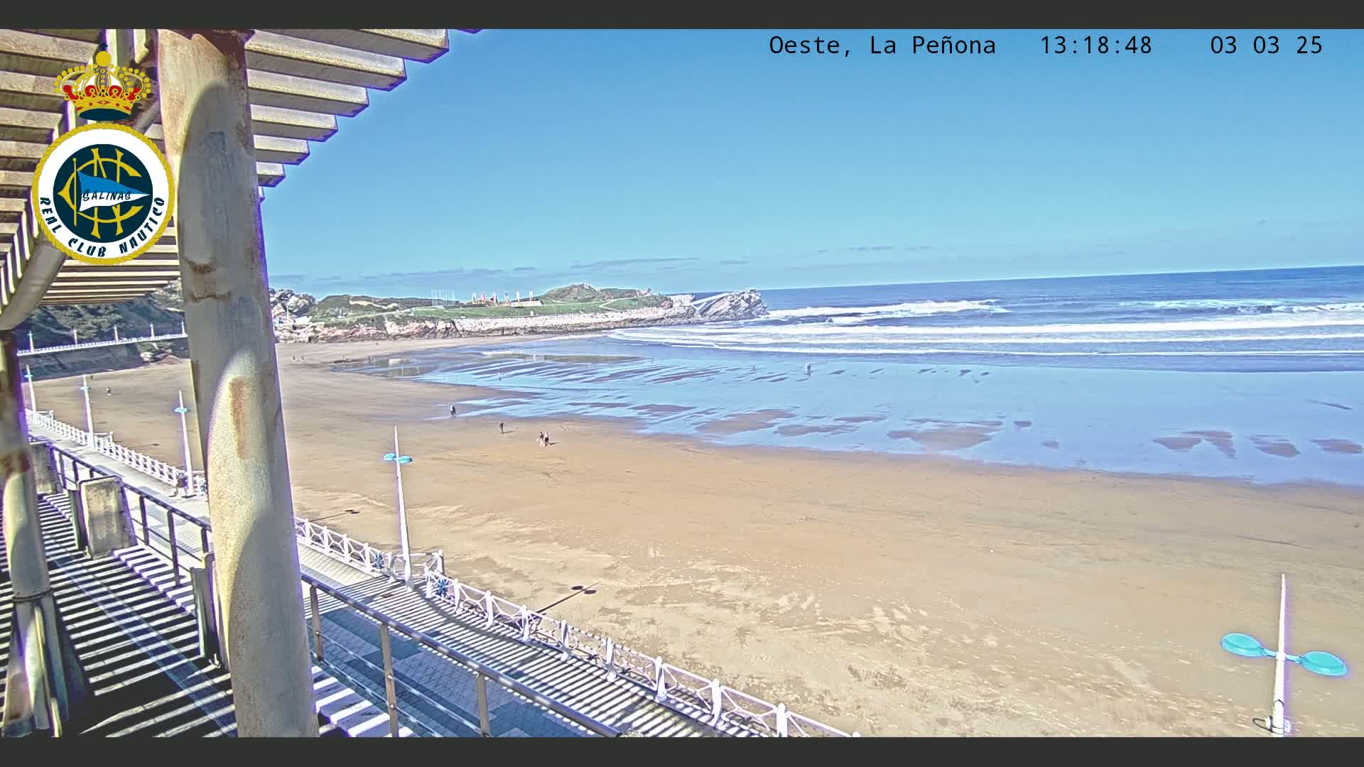 A sandy beach with gentle waves under a clear blue sky is viewed from a high vantage point on a sunny day.