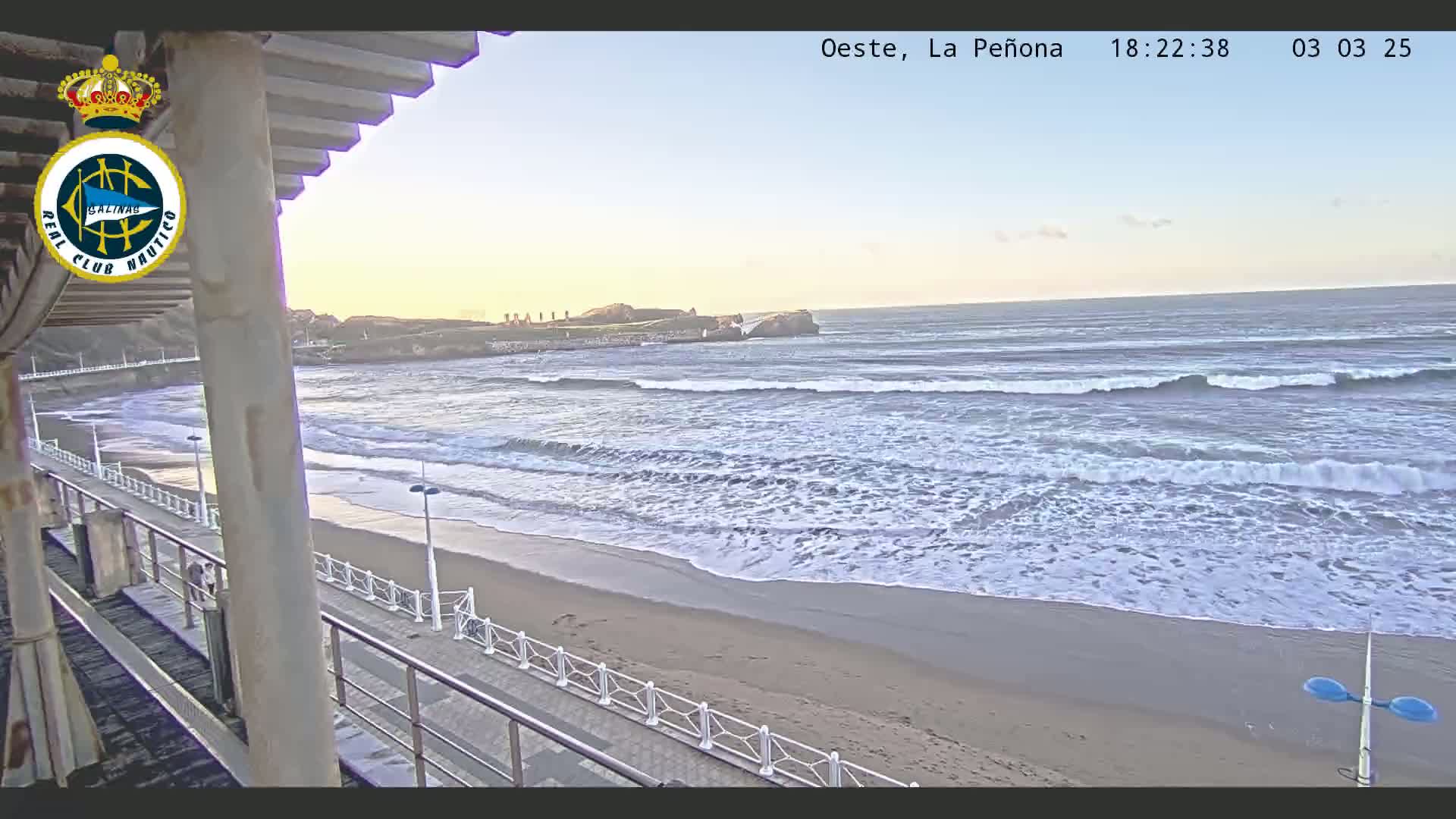 A sandy beach with moderate waves under a clear, pale sky is viewed from a seaside structure with a walkway.