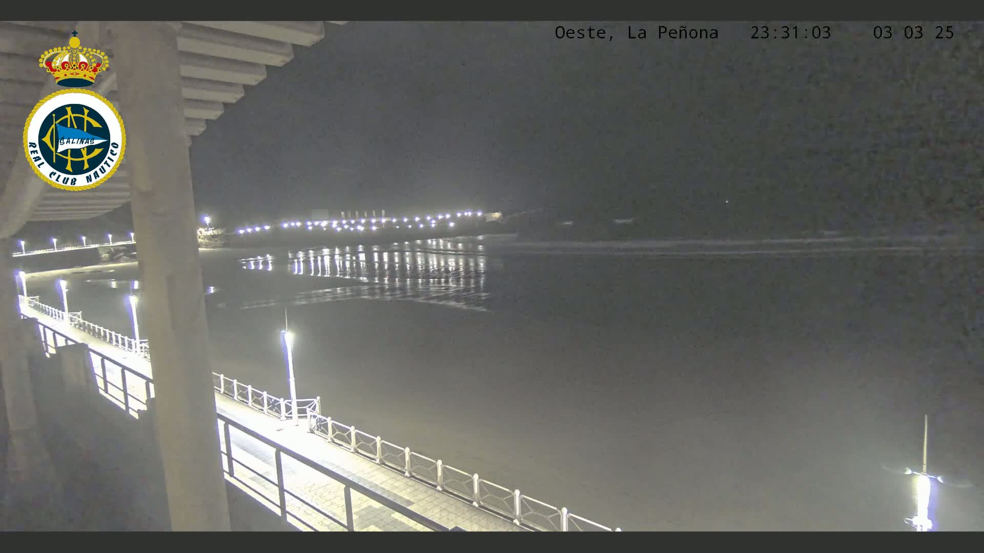 A nighttime view from a pier shows a calm, dark body of water reflecting lights from the shore, with gentle waves visible near the beach.