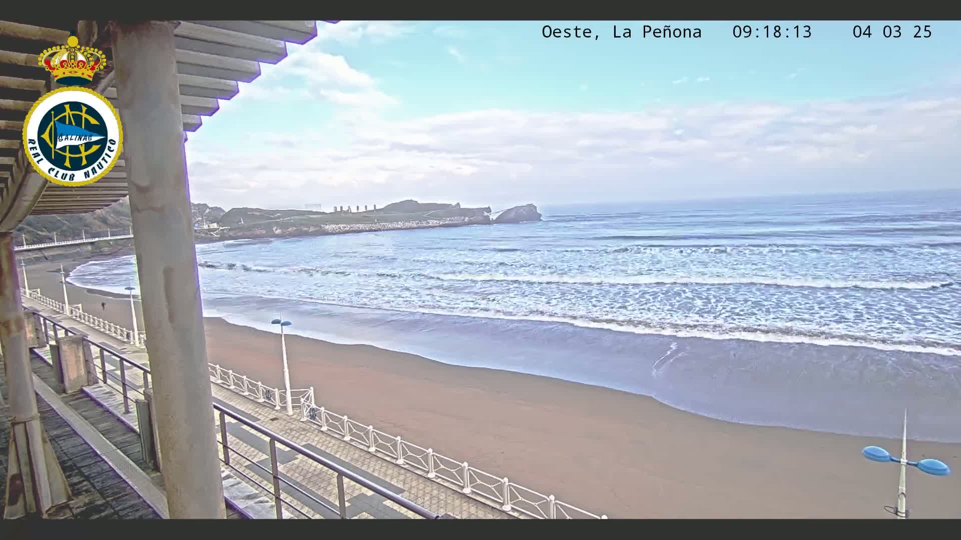 A sandy beach with gently rolling waves under a partly cloudy sky is viewed from a covered walkway.