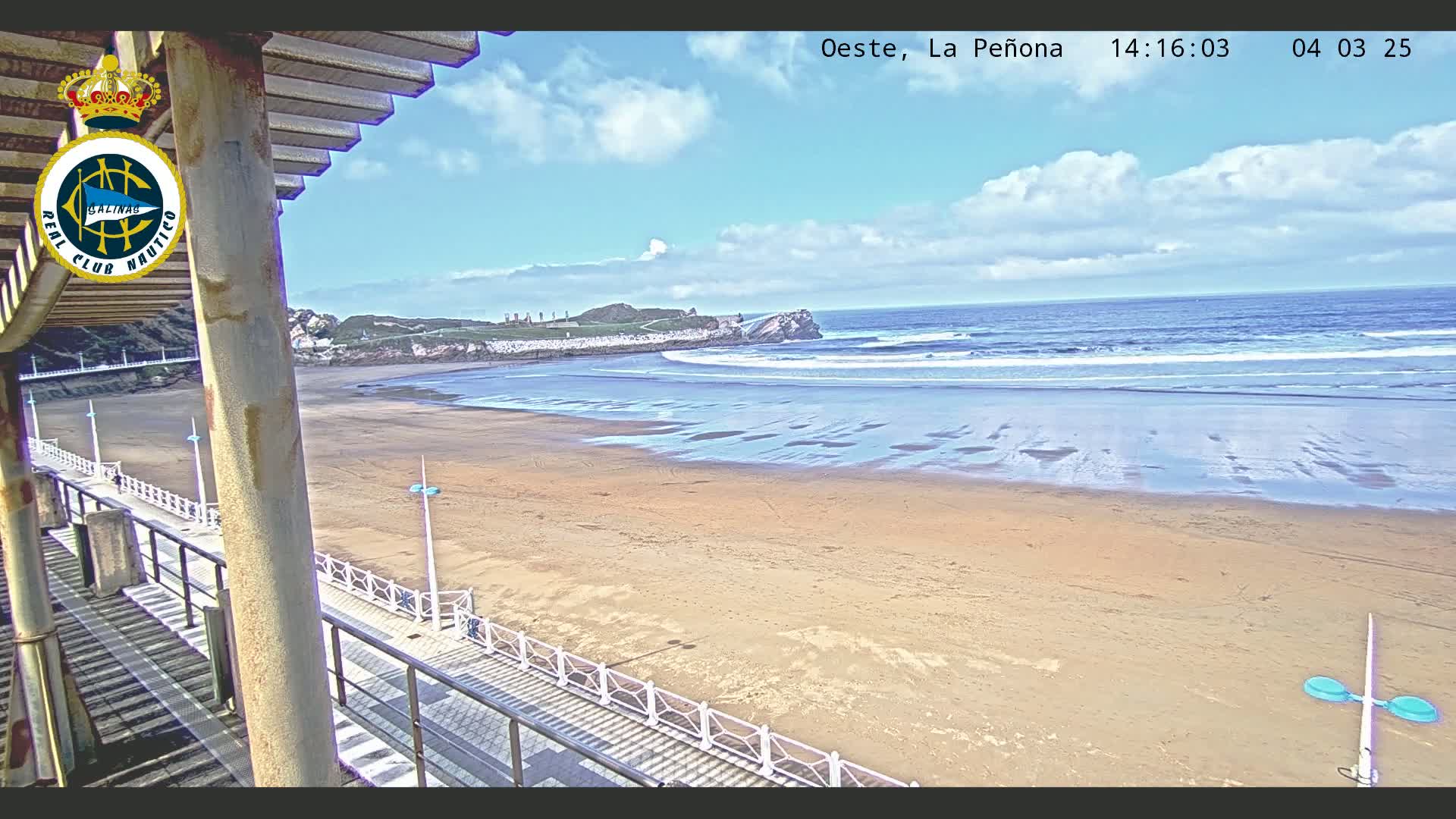 A sandy beach stretches out to a moderately wavy ocean under a partly cloudy sky, viewed from an elevated position on a walkway with railings.