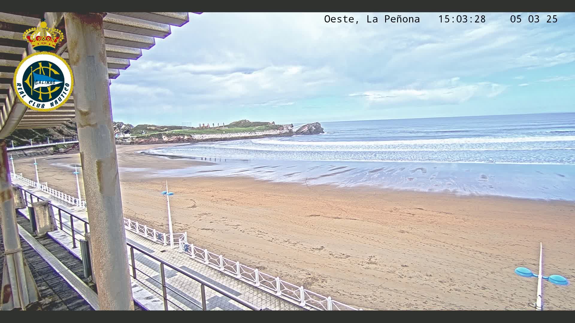 A sandy beach with gentle waves under a partly cloudy sky is viewed from a covered walkway.