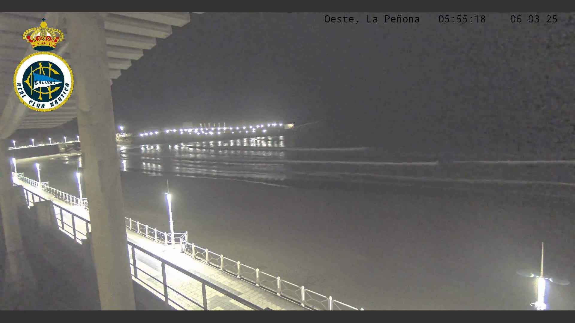 A nighttime view of a calm ocean with small waves lapping a shore at the end of a pier, illuminated by lights.