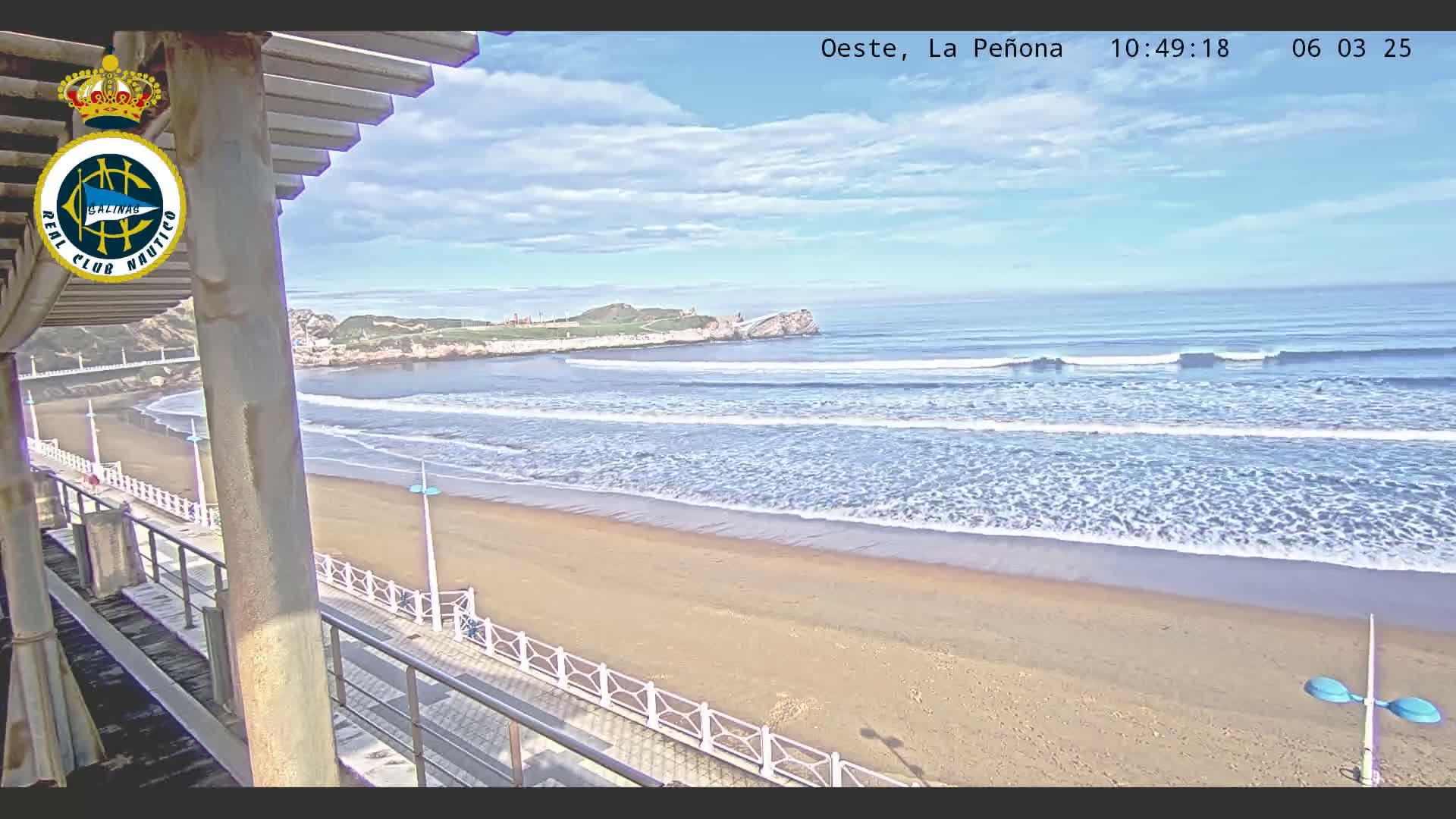 A sandy beach with gentle waves under a partly cloudy sky is viewed from an elevated position, showing a boardwalk and a distant headland.