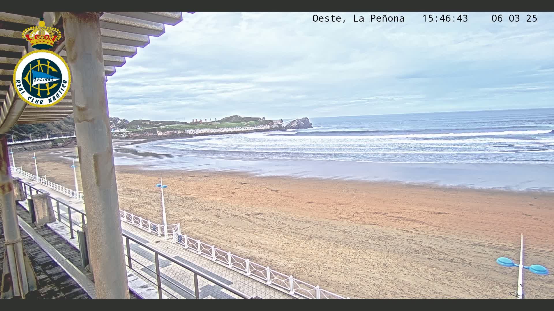 A sandy beach with gentle waves under a cloudy sky is viewed from an elevated position, showing a boardwalk leading towards the ocean.