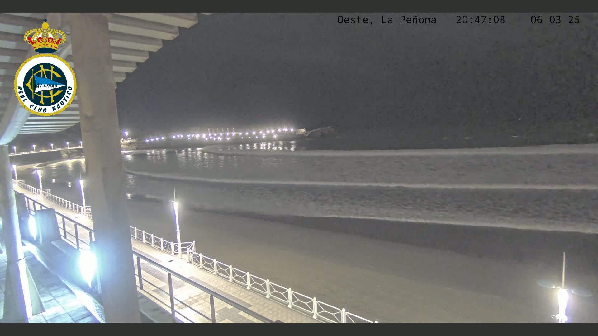 A nighttime view of a beach with gentle waves lapping the shore, illuminated by lights along a boardwalk.
