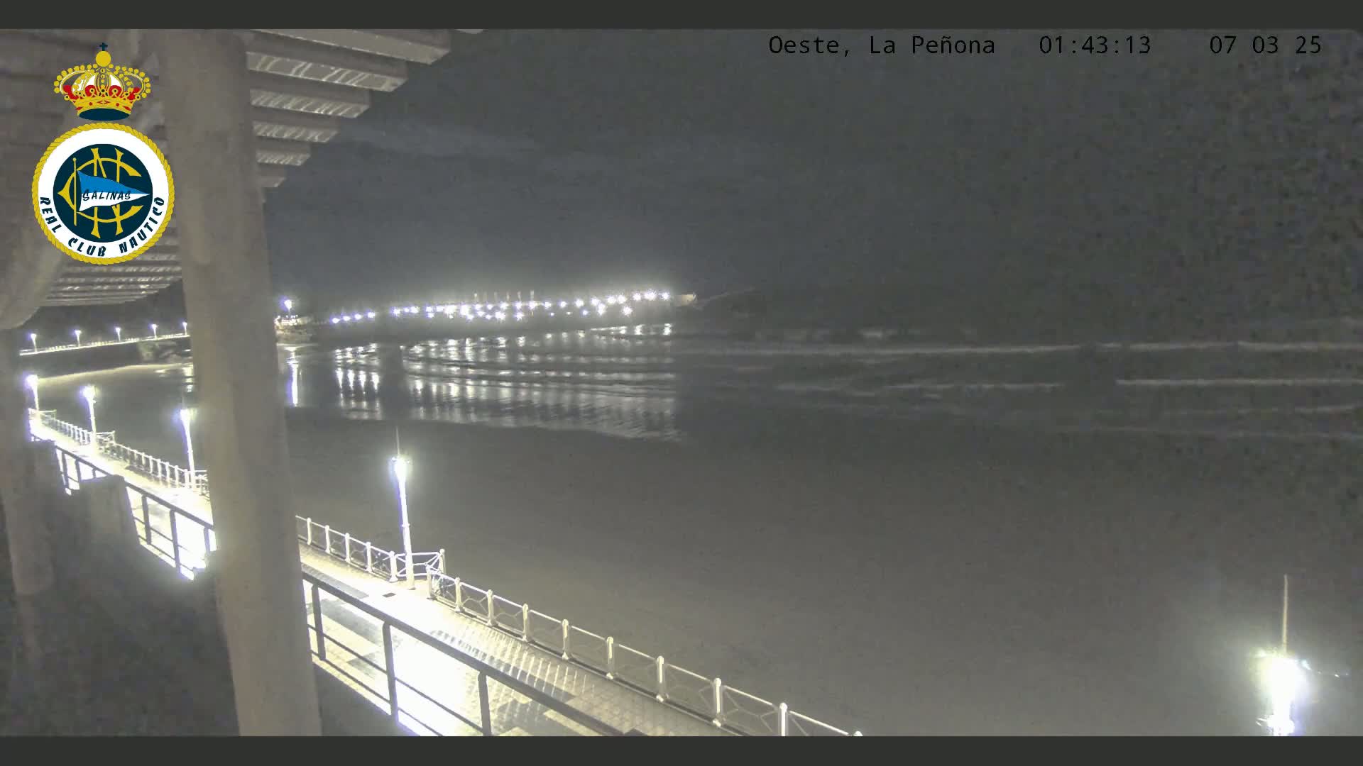 A nighttime view from a pier shows a dark, calm ocean with gentle waves lapping the shore, illuminated by lights along the pier and in the distance.