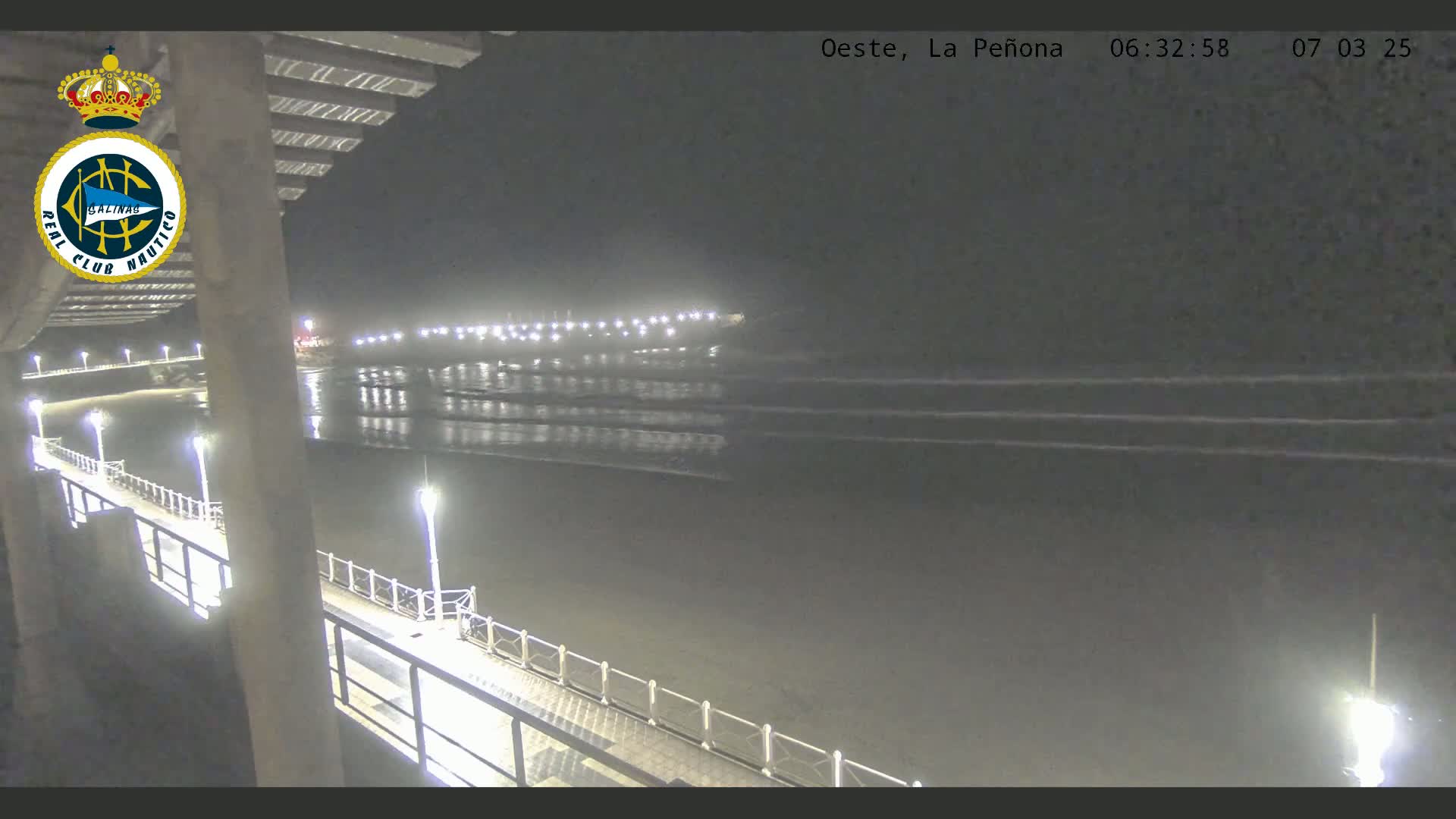 A nighttime view from a pier shows a calm, dark ocean with gentle waves lapping at the shore, under a clear sky.