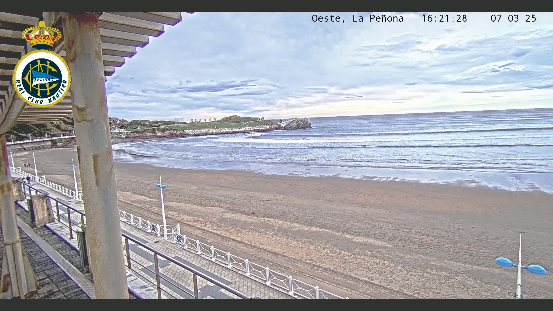 A sandy beach with gentle waves rolls onto the shore under a partly cloudy sky, seen from an elevated vantage point overlooking a boardwalk.