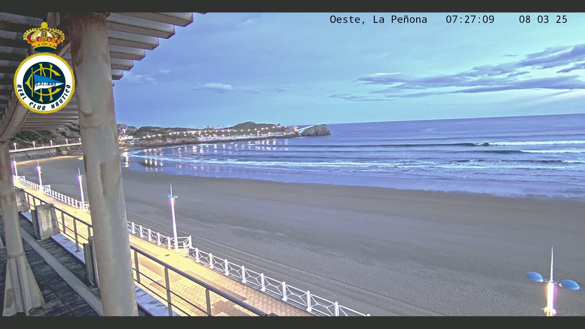A sandy beach at dawn, under a partly cloudy sky, features gentle waves rolling toward the shore, a boardwalk with lights, and a rocky outcrop in the distance.