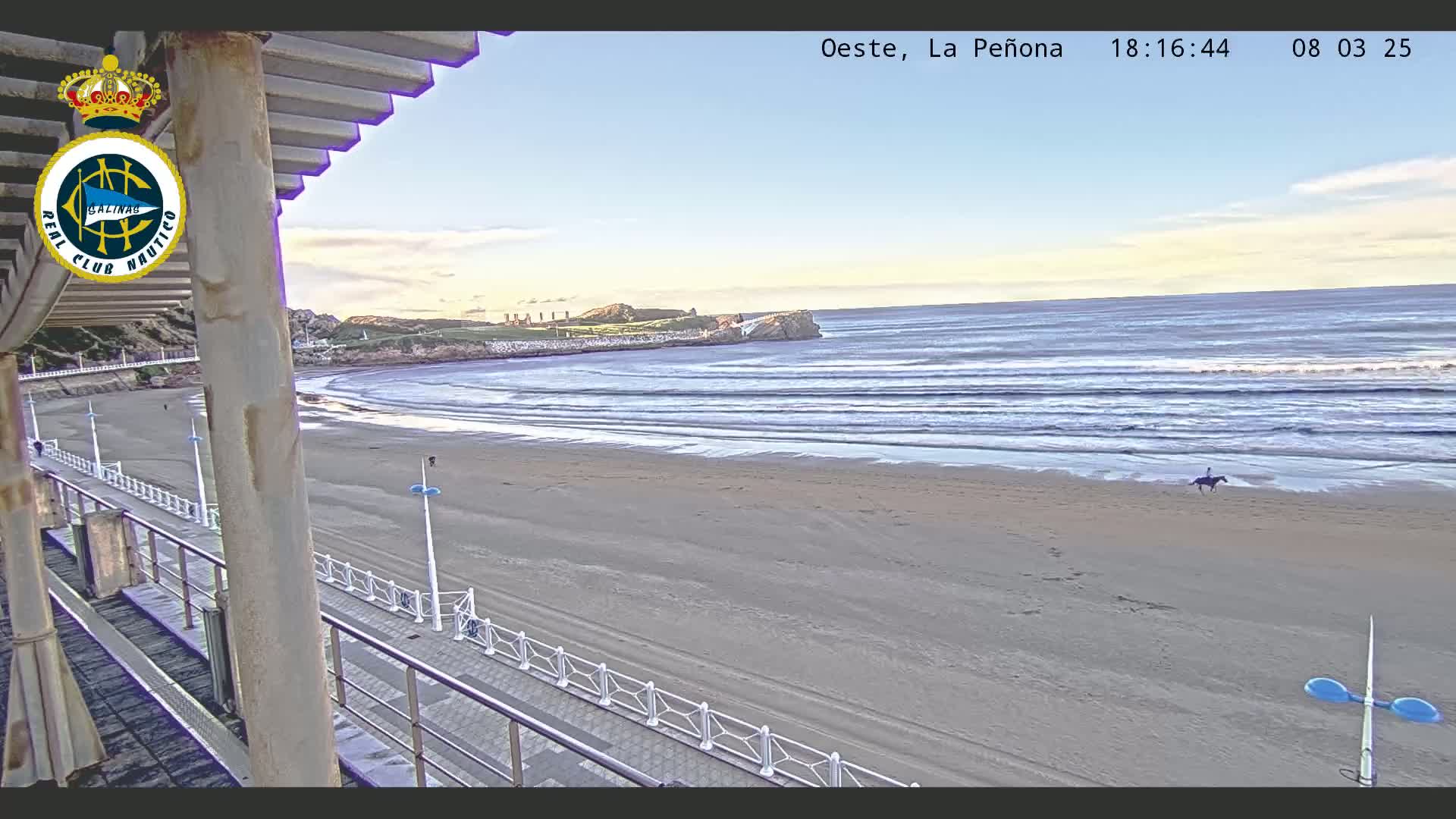 A sandy beach with gentle waves under a clear, light blue sky is seen from a boardwalk with a person on horseback in the distance.