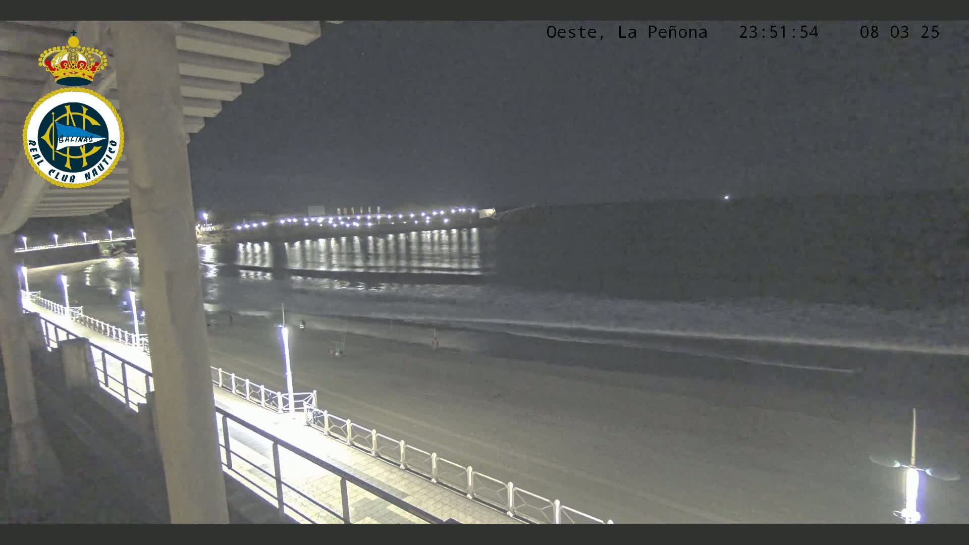 A nighttime view from an elevated position shows a beach with gentle waves lapping the shore under a clear, dark sky;  a pier with lights extends into the water.