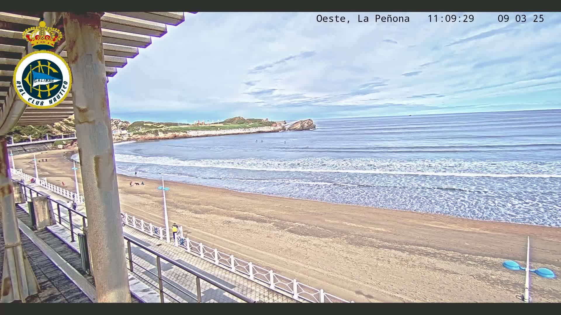 A sandy beach with gentle waves rolls into the ocean under a partly cloudy sky, viewed from a covered balcony.
