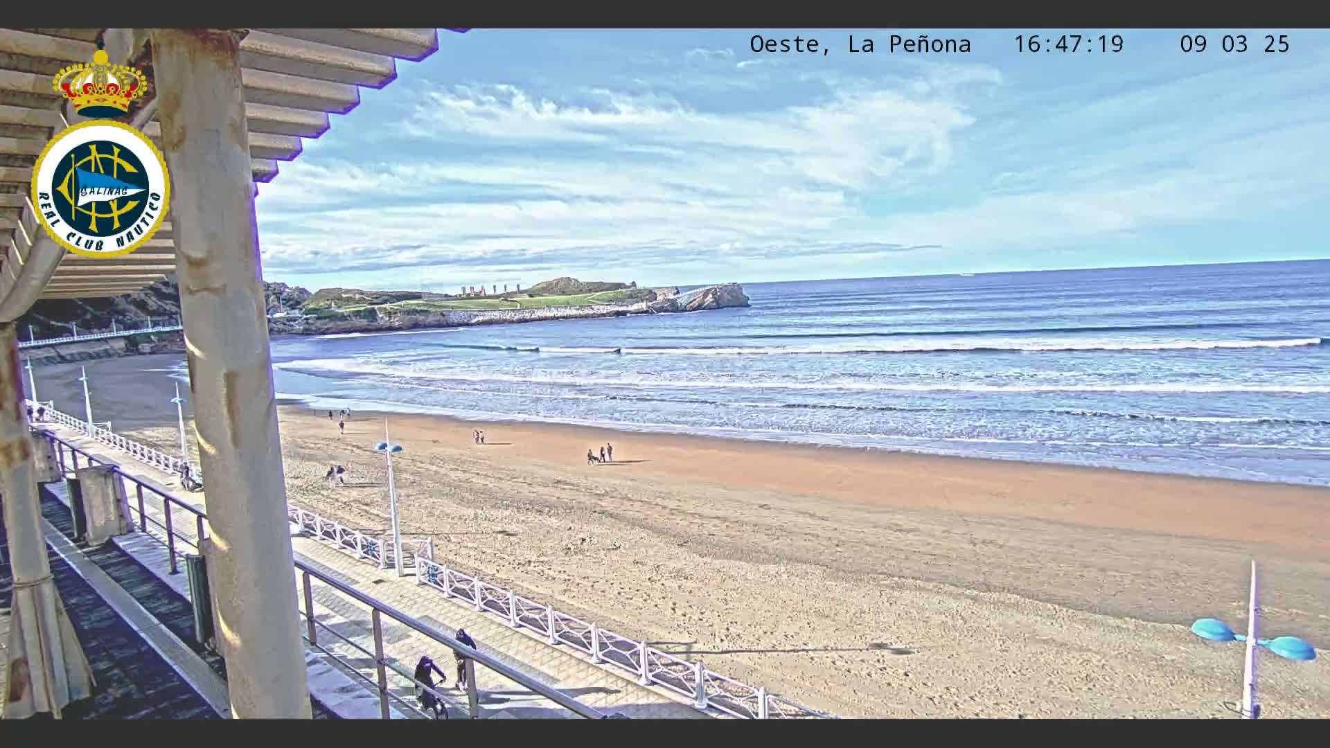 A sandy beach with a few people walking along the shore, under a partly cloudy sky, with gentle waves rolling into the ocean.