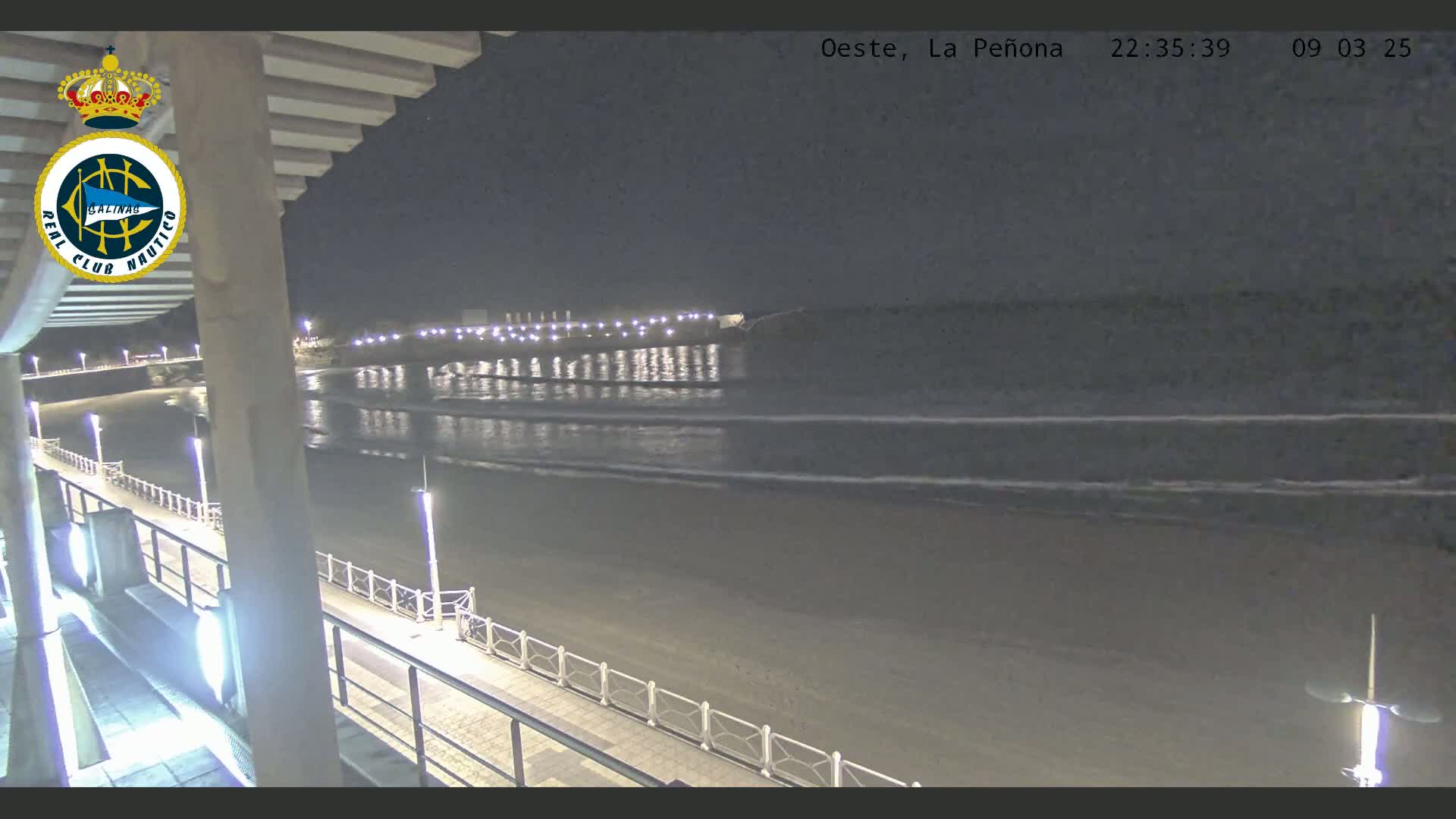 A nighttime view of a beach with calm waves gently lapping the shore, illuminated by lights along a boardwalk.