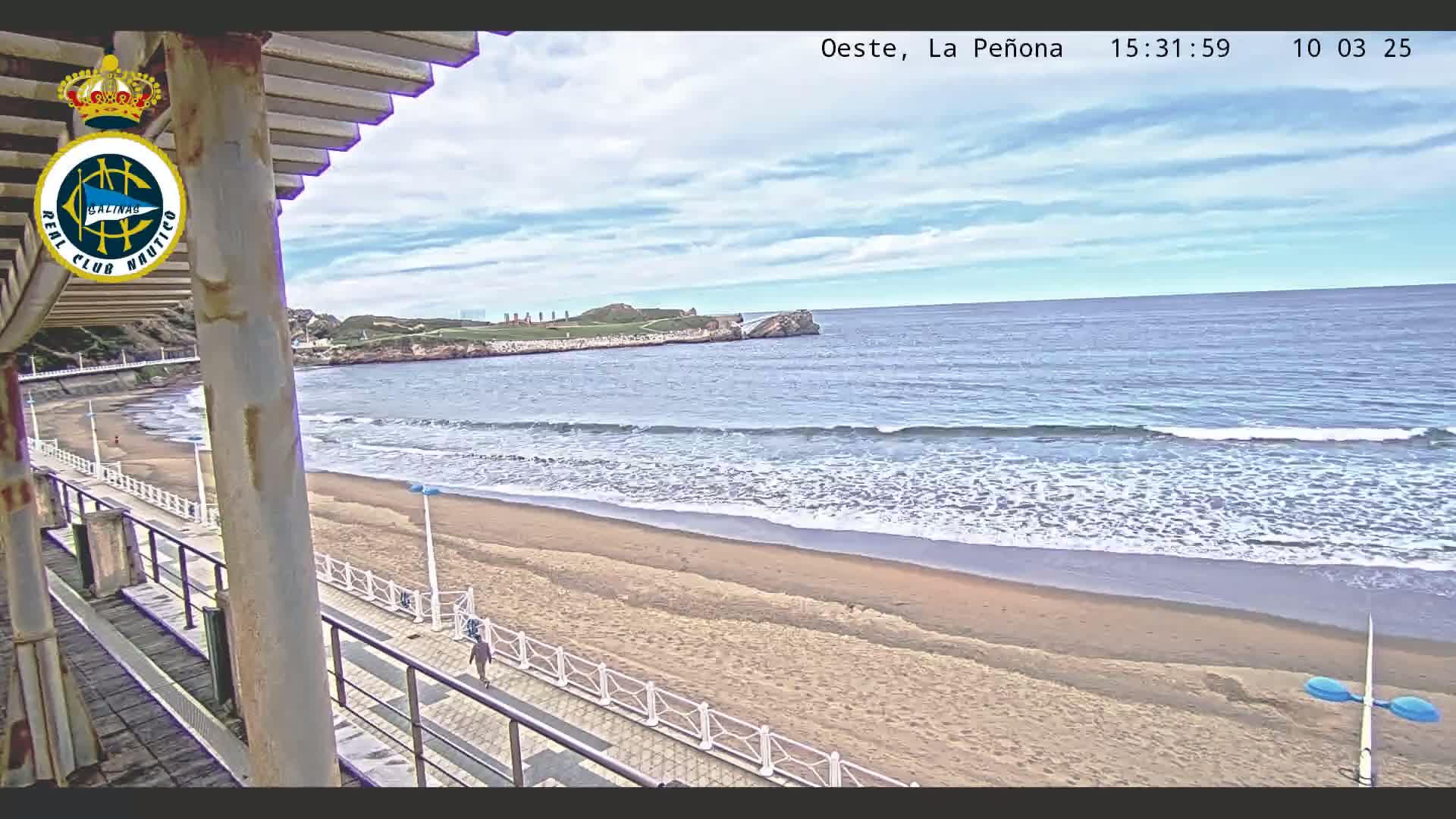 A sandy beach with gentle waves laps at the shore under a partly cloudy sky, viewed from an elevated vantage point overlooking a boardwalk.