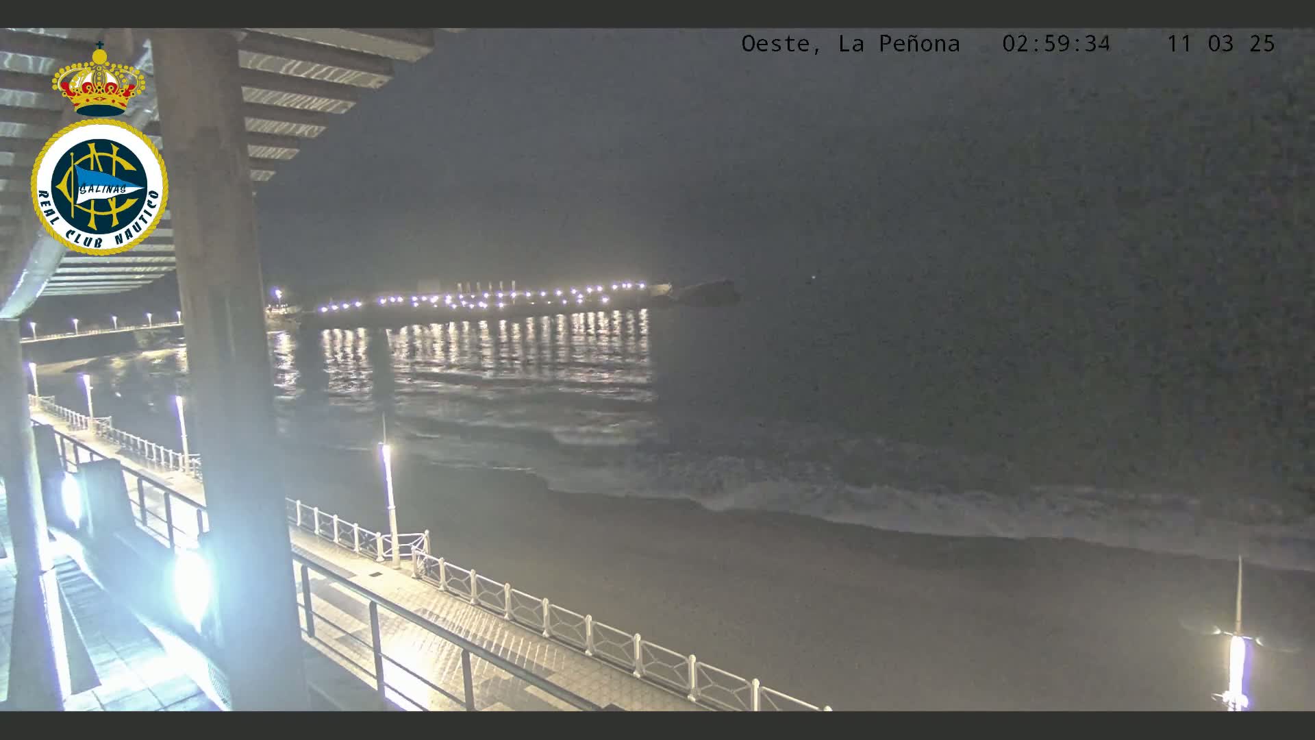 A nighttime view of a dark ocean with gentle waves lapping a shoreline illuminated by lights, seen from an elevated position overlooking a boardwalk.