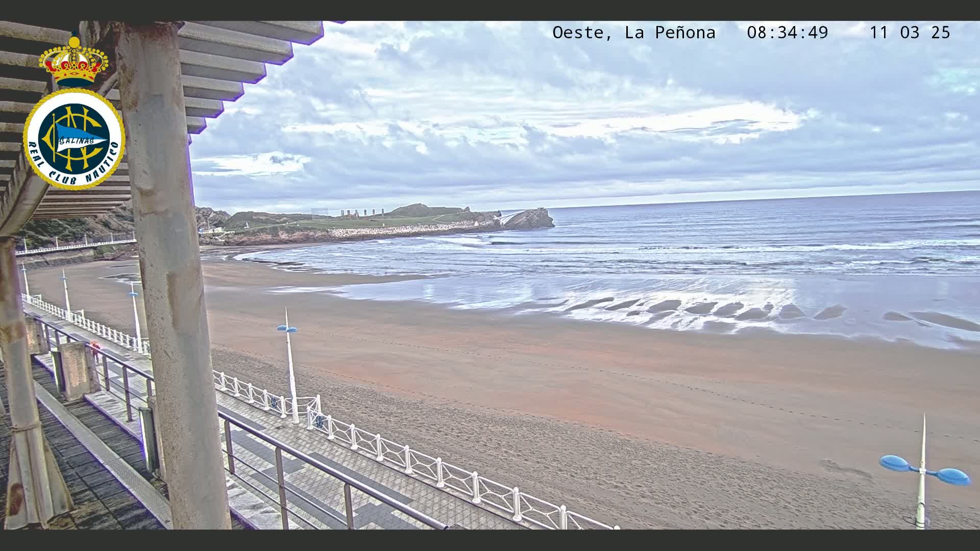A sandy beach with gentle waves under a cloudy sky is viewed from a covered walkway.