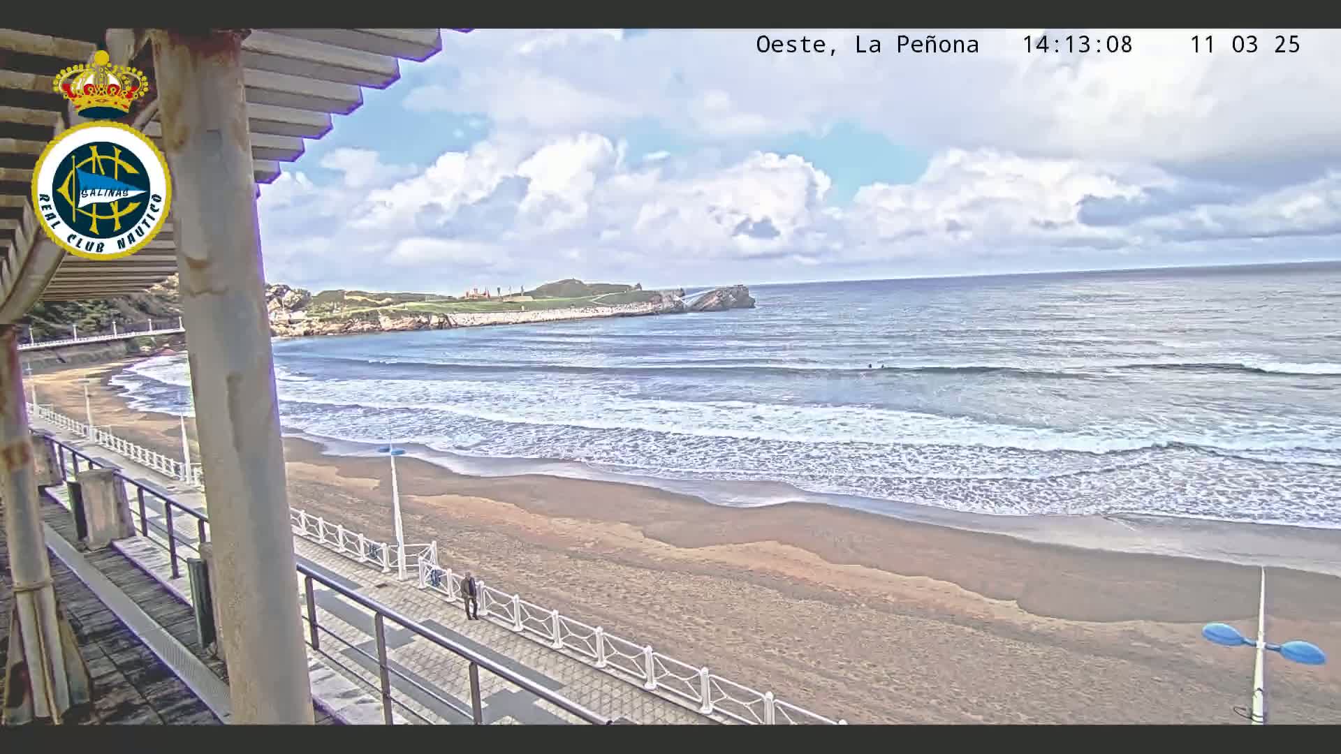A partly cloudy day shows a sandy beach with gentle waves lapping the shore, a boardwalk with a few people walking, and a rocky point in the distance.