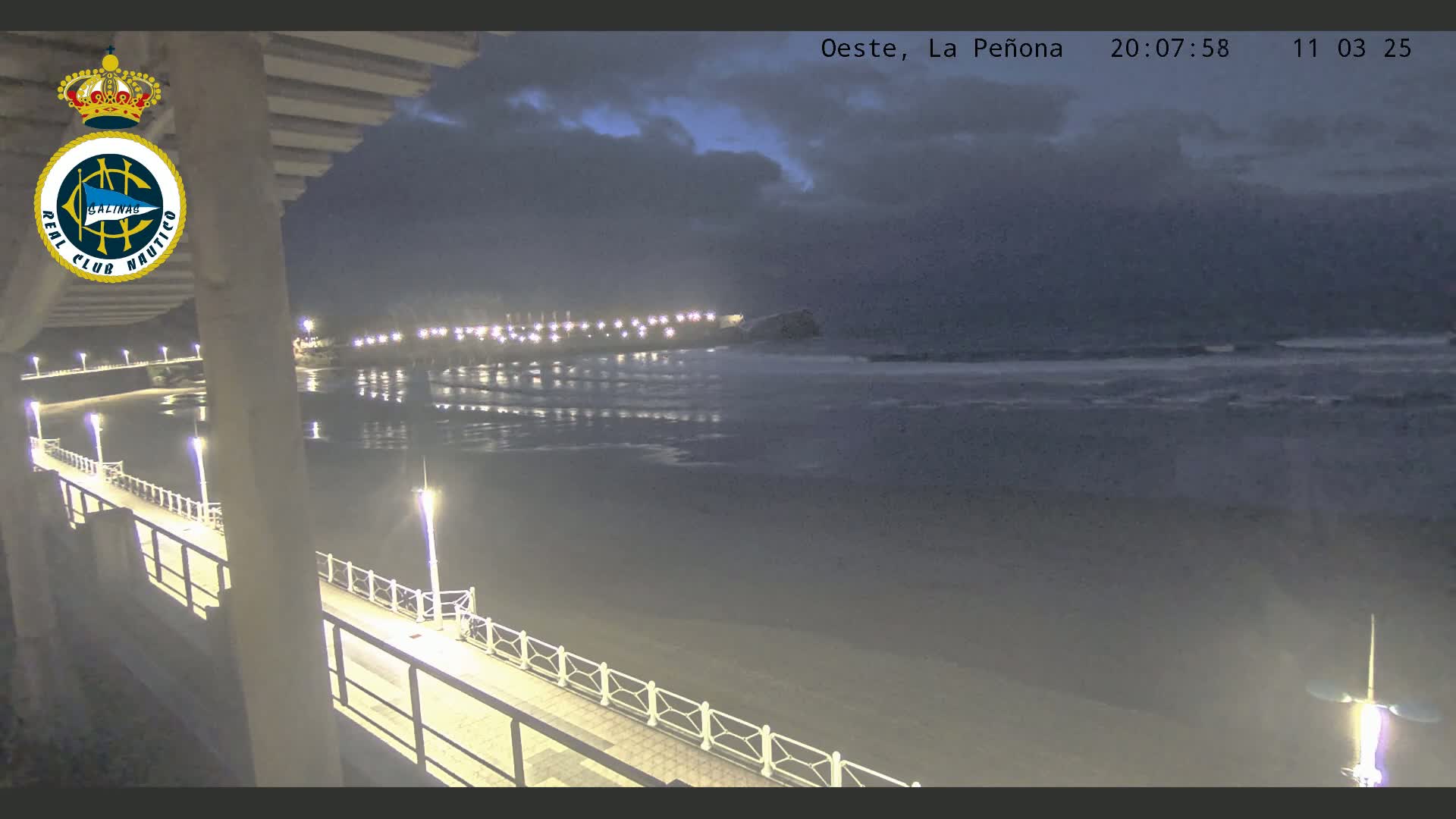 A nighttime view of a calm ocean, a beach with lights, and a pier extending from a building under a cloudy sky.