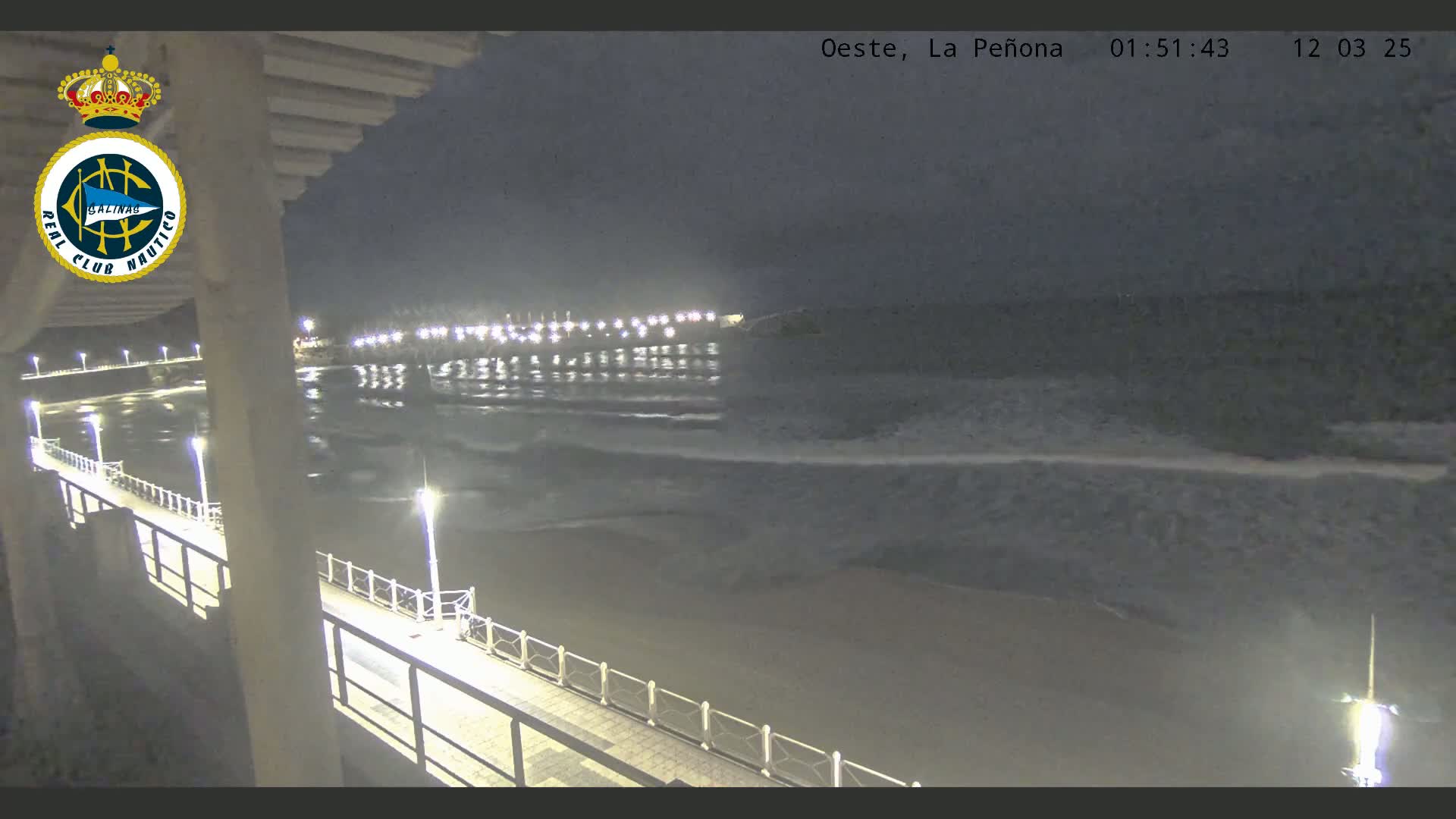 A nighttime view from a pier shows a dark beach and ocean with small waves under a cloudy sky, illuminated by lights along the pier and in the distance.