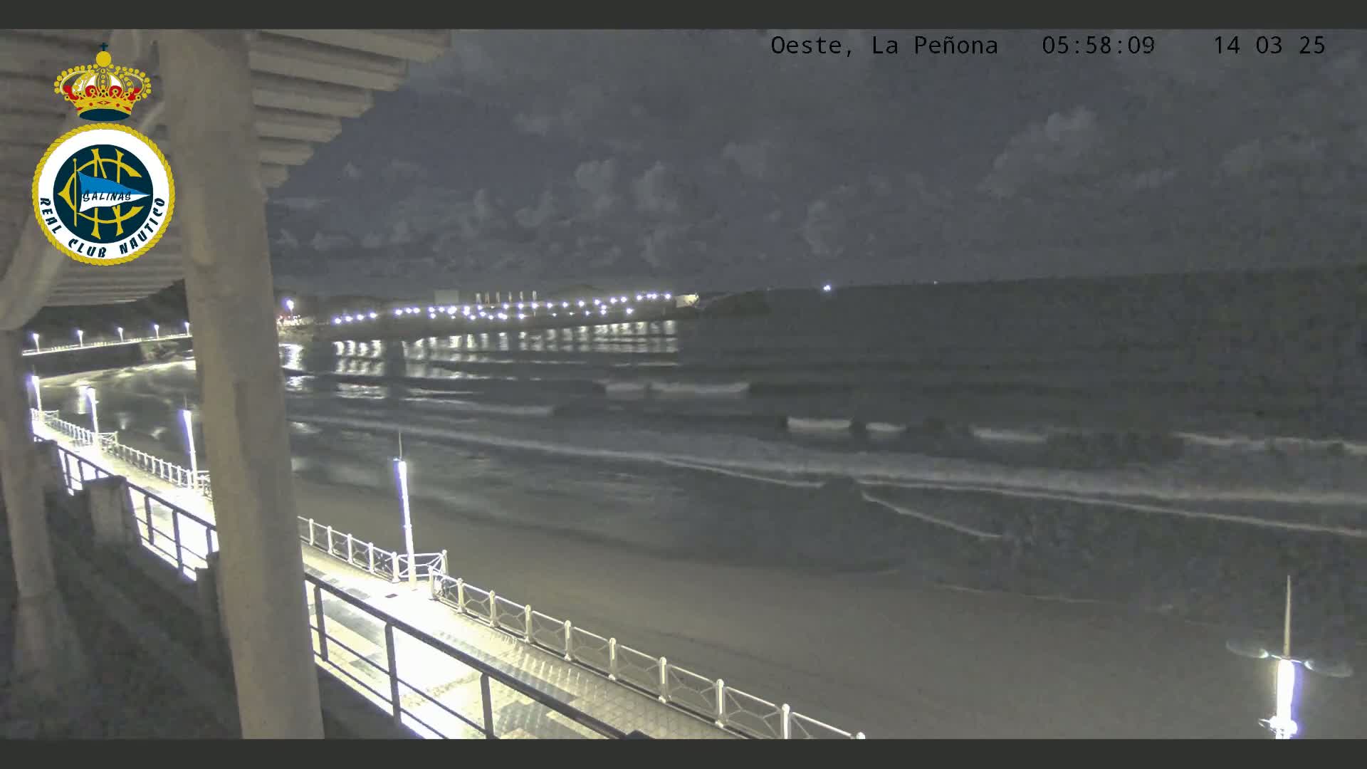 A nighttime view from an elevated position shows a beach with gently rolling waves under a partly cloudy sky, and a pier extending into the water with lights illuminating the walkway.