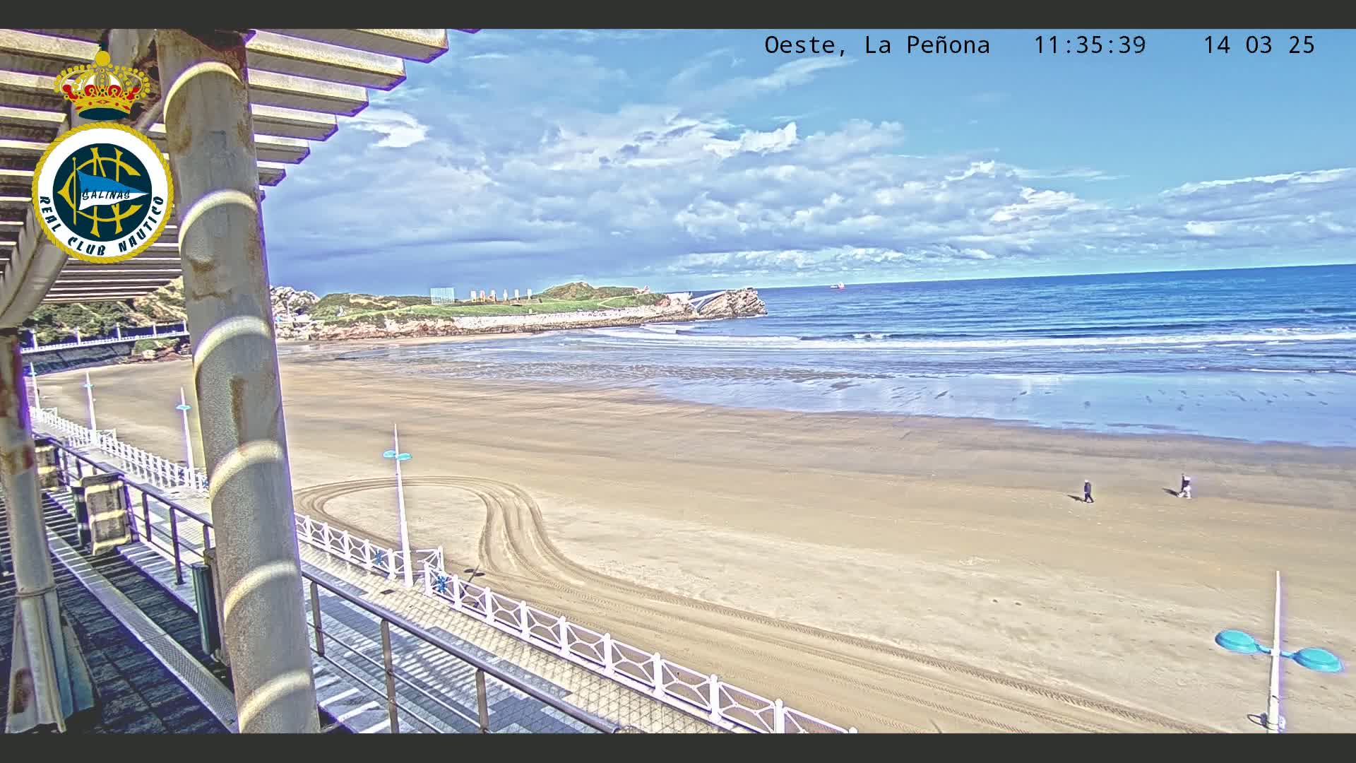 A sandy beach with a few people walking stretches out to a wavy ocean under a partly cloudy sky.
