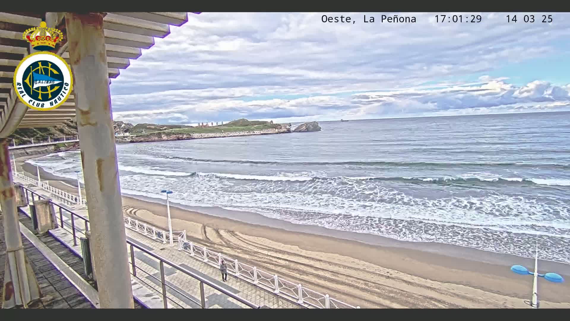 A sandy beach with gentle waves under a partly cloudy sky is viewed from an elevated position, overlooking a boardwalk and a rocky outcrop in the distance.