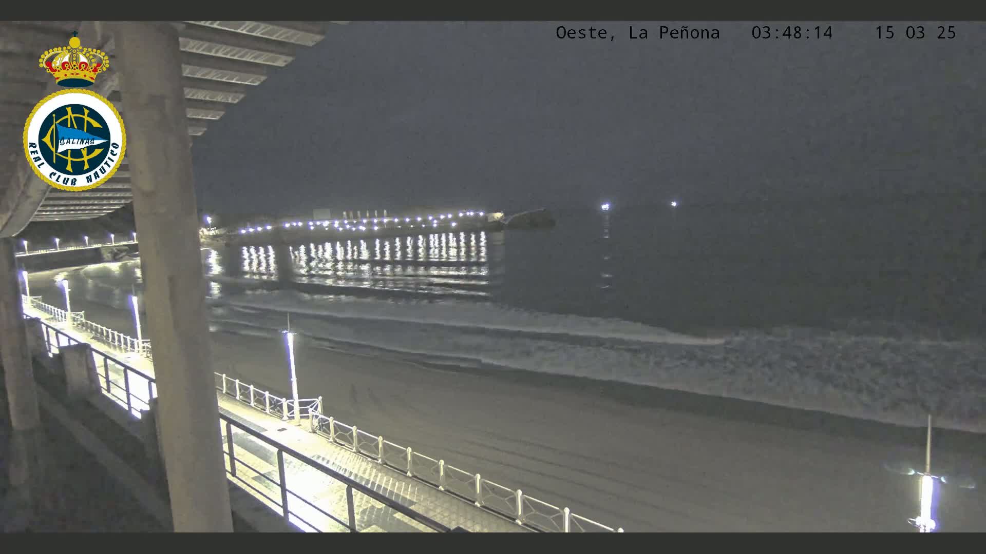A nighttime view from an elevated position shows a sandy beach, gentle waves lapping the shore, and a pier extending into calm waters under a dark sky.