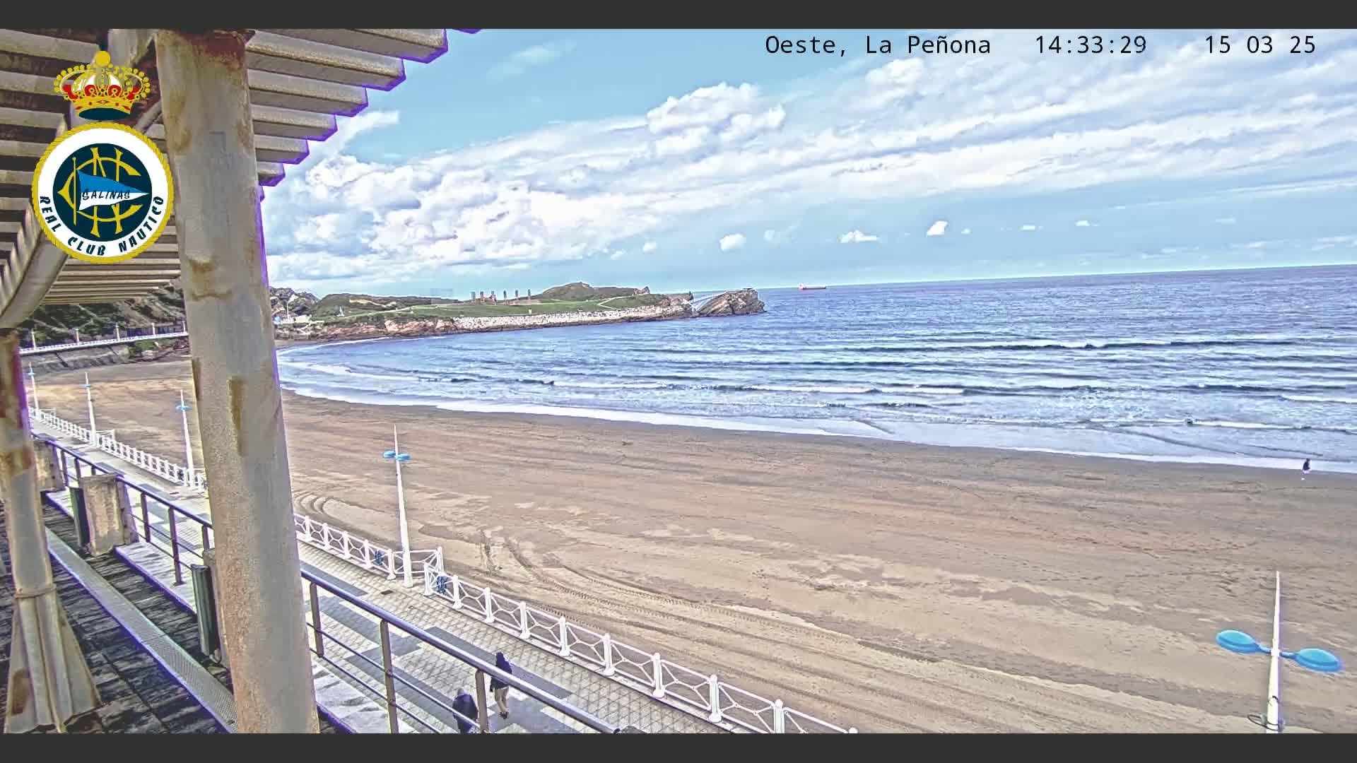 A sandy beach with small waves under a partly cloudy sky is viewed from an elevated vantage point, showing a boardwalk and a few people walking along the shore.