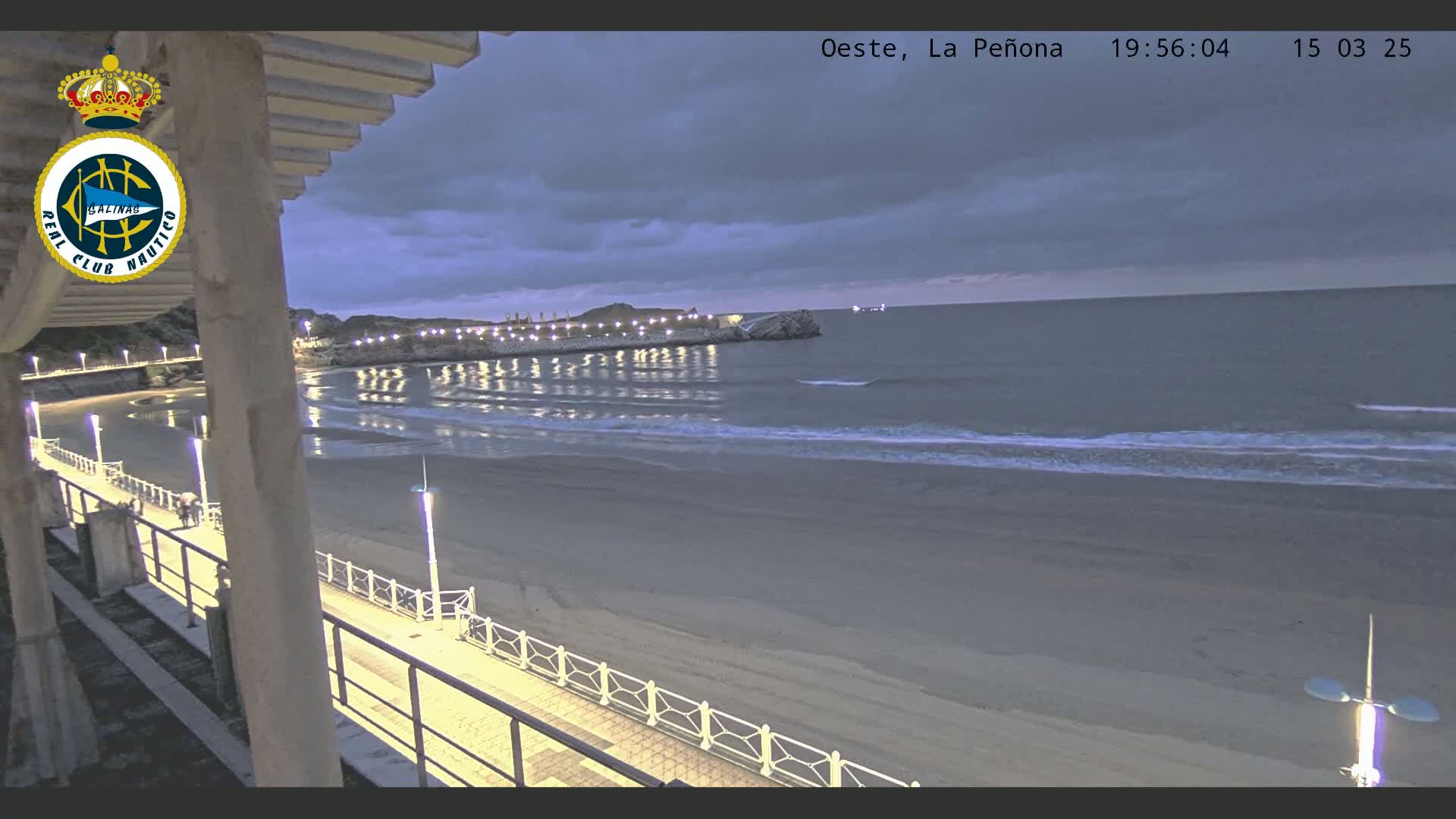 A nighttime view from an elevated vantage point shows a sandy beach, calm ocean waves, a lit walkway along the shore, and a distant rocky outcrop under a cloudy sky.