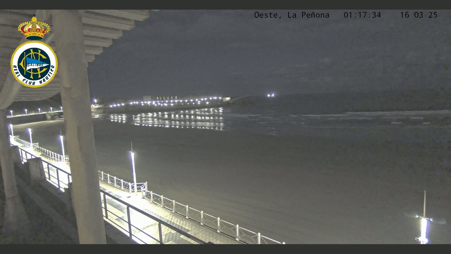 A nighttime view from a pier shows a mostly calm, dark ocean, a lit beach, and a distant shoreline.