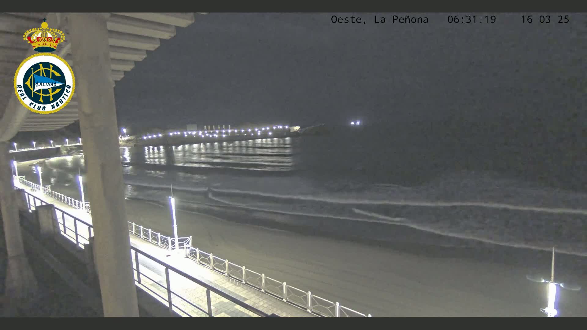 A nighttime view from a pier shows a sandy beach with gentle waves lapping the shore under a dark sky.