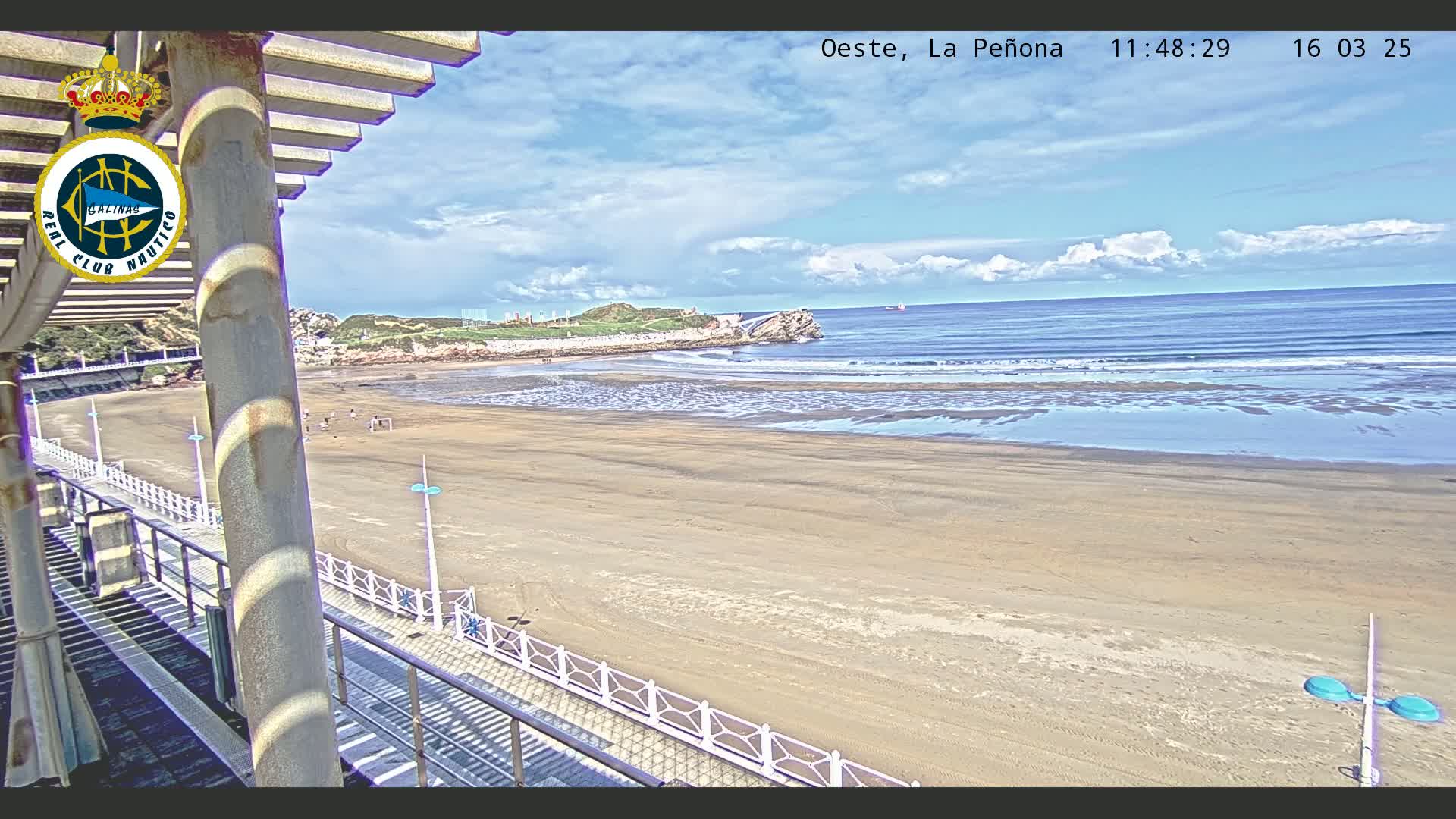 A sandy beach with gentle waves under a partly cloudy sky is seen from an elevated viewpoint, overlooking a boardwalk and a structure with a shaded area.