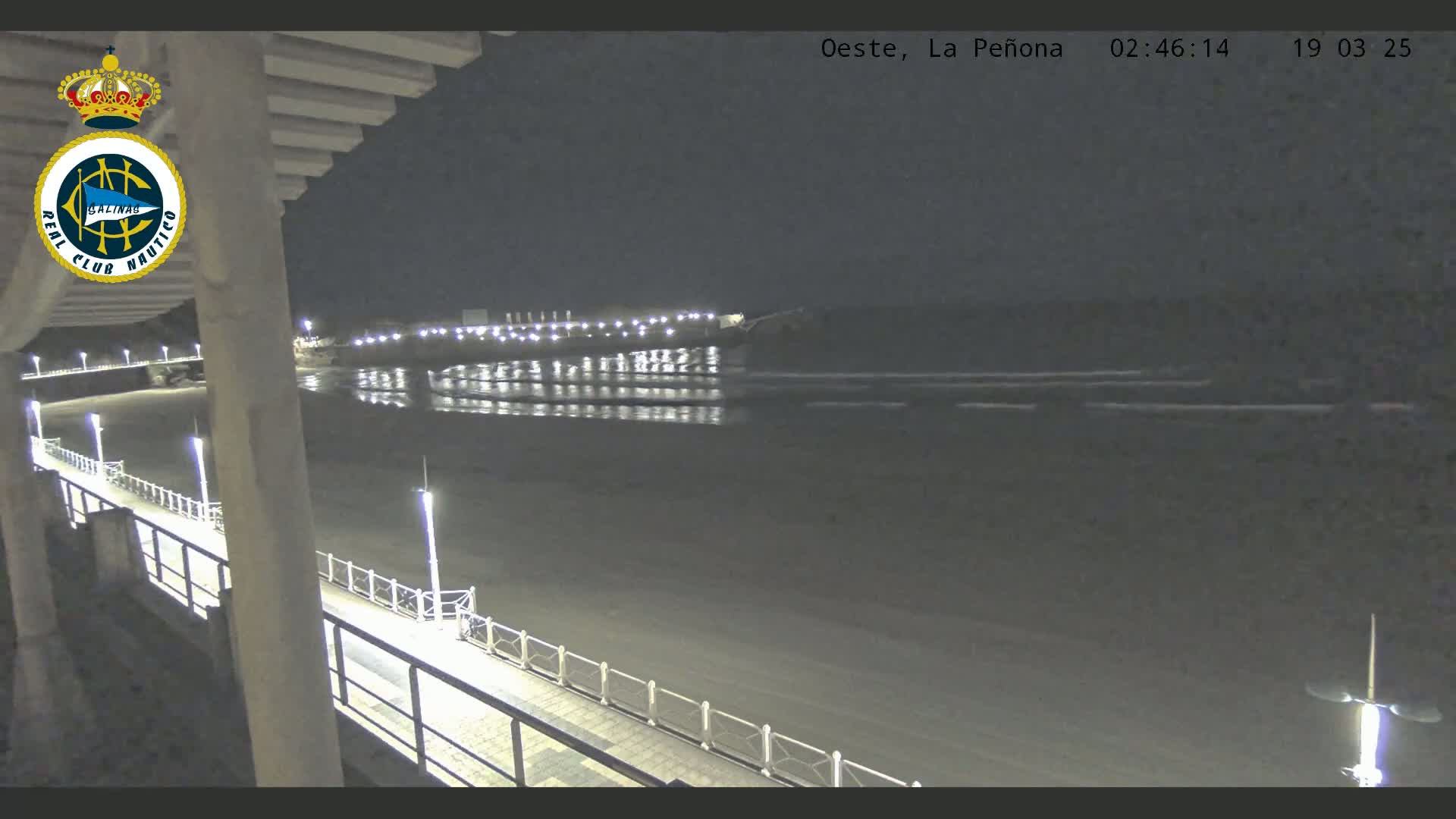 A nighttime view from an elevated position shows a long, lit walkway beside a dark beach and calm water reflecting lights from a distant structure.