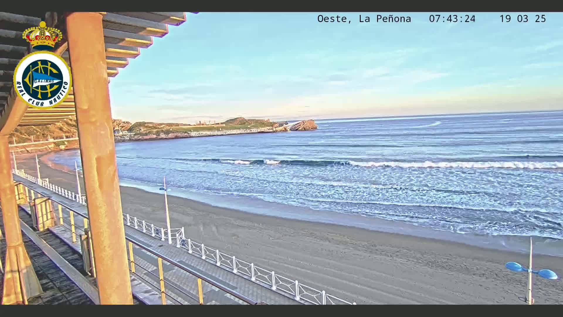 A sandy beach with gentle waves laps the shore under a clear sky, viewed from an elevated position on a boardwalk with a wooden structure overhead.