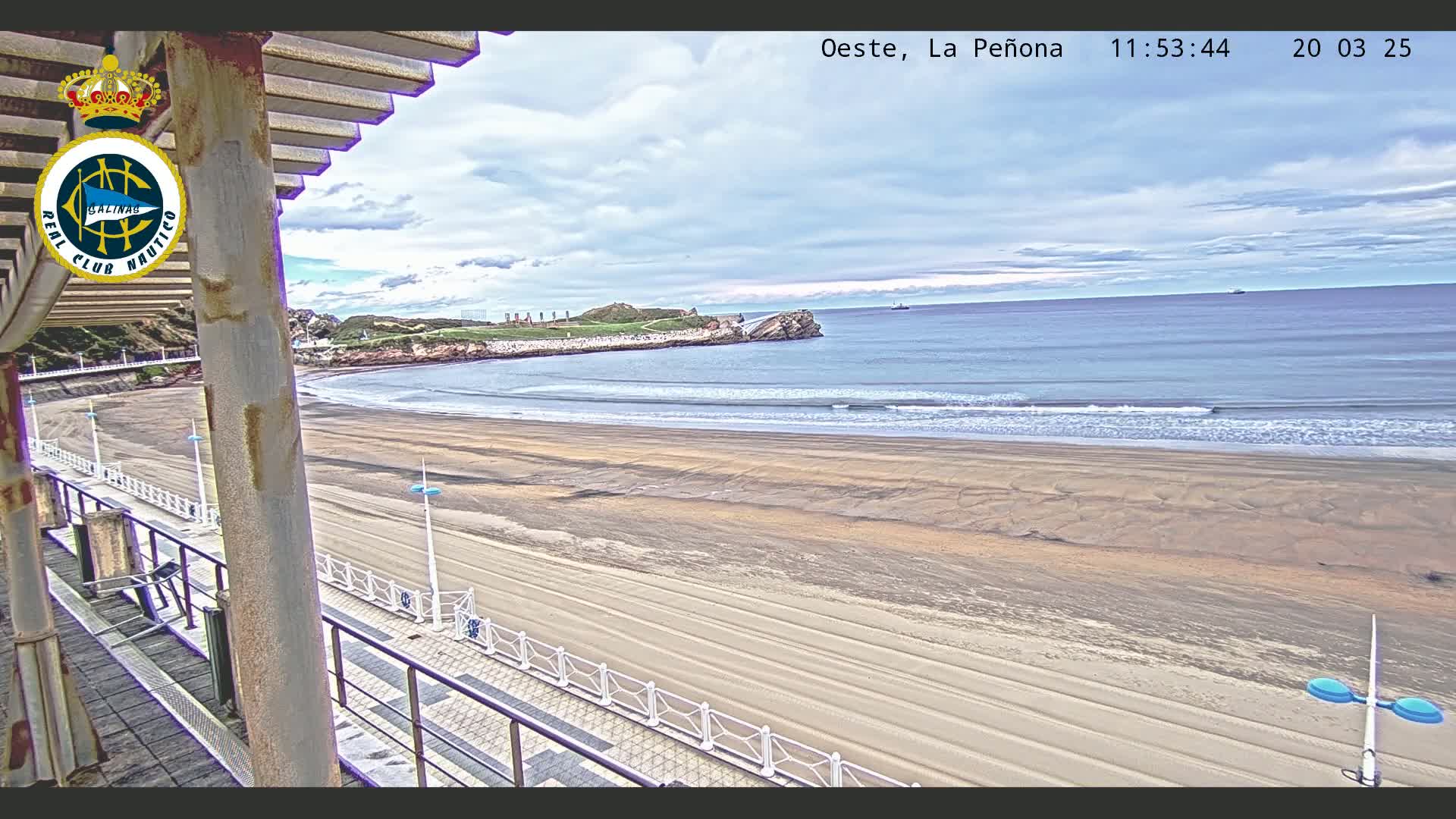 A sandy beach stretches out to a calm ocean under a partly cloudy sky, viewed from an elevated position on a boardwalk with railings.