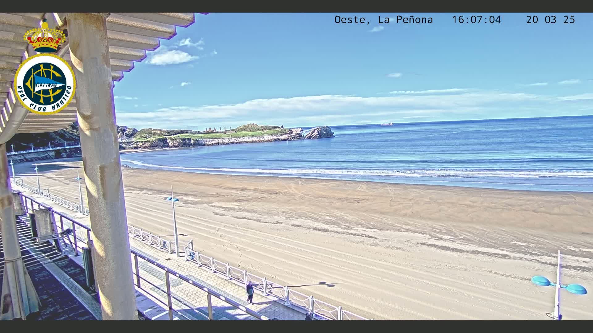 A sandy beach stretches to a calm ocean under a mostly sunny sky, viewed from a structure with a railing and a walkway.