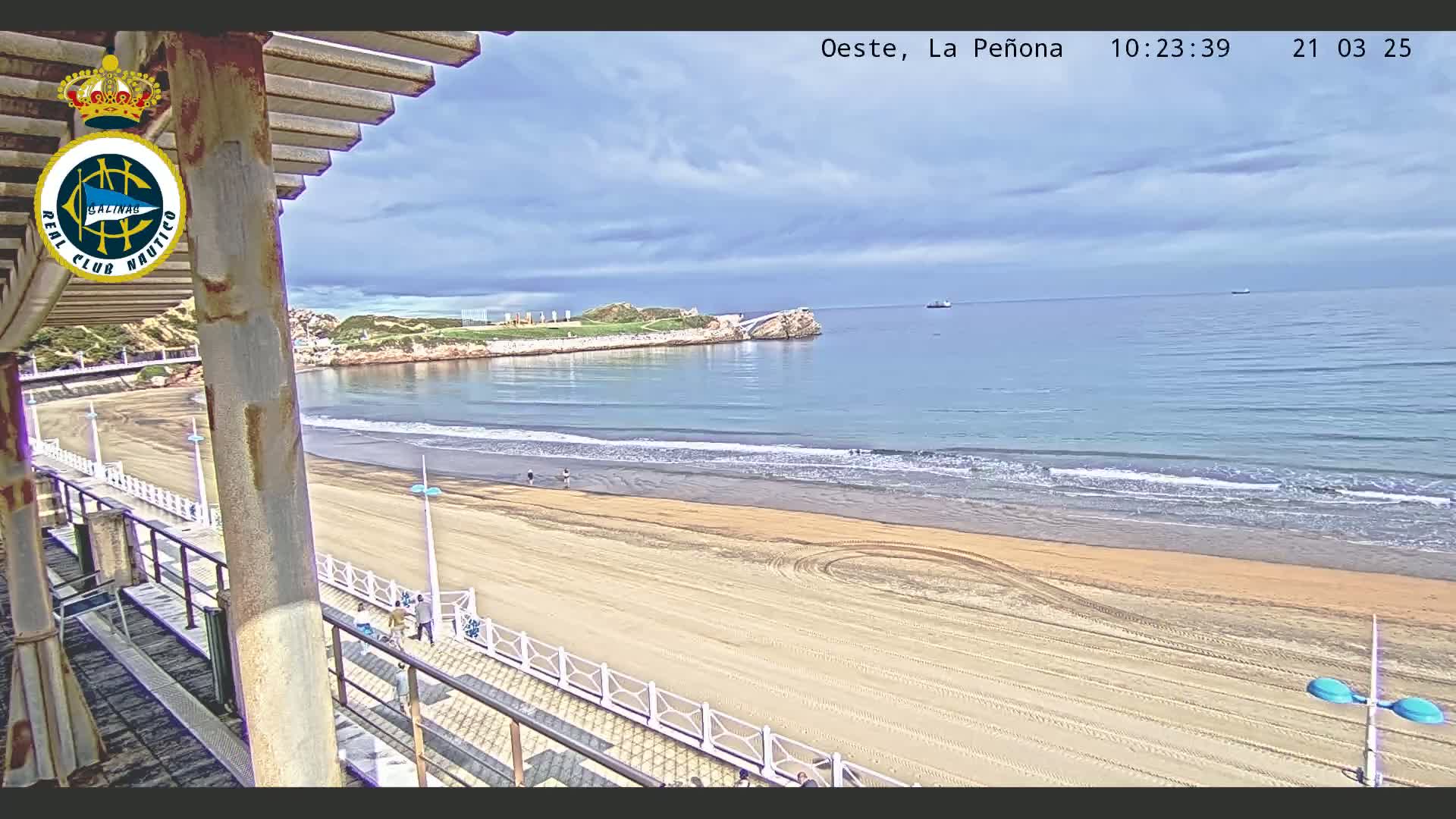 A sandy beach with gentle waves under a partly cloudy sky is viewed from an elevated position, showing a boardwalk and a portion of a building with a pergola.