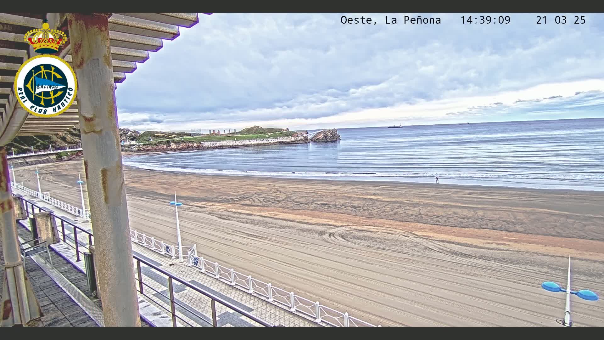 A sandy beach stretches out to a calm ocean under a cloudy sky, viewed from a covered walkway.